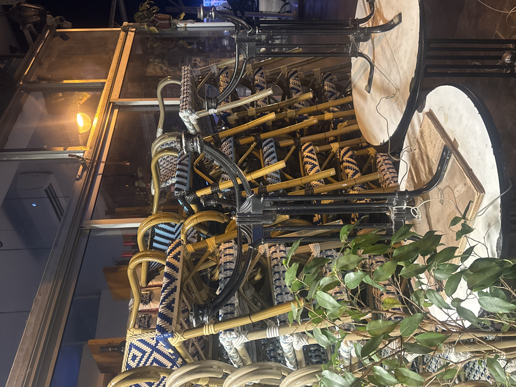 Stacked furniture, including chairs with blue and white woven patterns, along with round tables with black metal frames, are observed. Some plant foliage is present in the lower right. The location is suggested to be Ramat Gan, Israel.