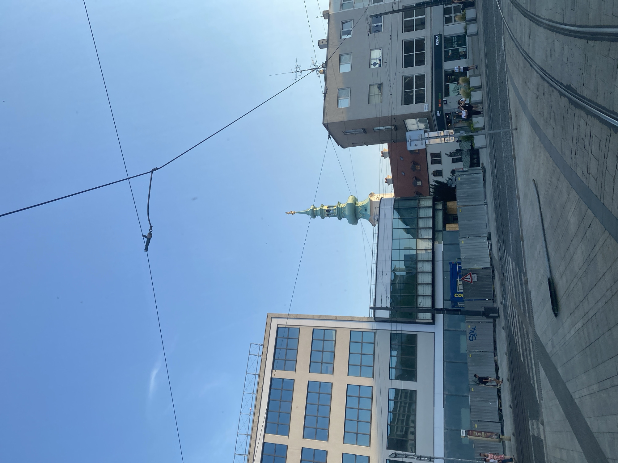 This media file depicts an outdoor scene in Bratislava, Slovakia, with a clear blue sky indicating daytime, likely a sunny day. The foreground features tram tracks running alongside a paved walkway. In the mid-ground, several buildings are visible. To the left, a modern building with a glass facade and multiple windows stands out. Further in the background, a historical building with a distinctive green spire topped with a golden element is prominent, suggesting a church or cathedral. Adjacent to it, a grey, multi-story building with numerous windows is also present. A few people are visible on the walkway, some standing in small groups, others walking, suggesting a normal day with pedestrian activity. Overhead, power lines crisscross the sky, likely for the trams. Construction fencing and materials are also visible in the mid-ground, indicating ongoing development or renovation. Text visible on the buildings includes "REGINA" and "COFFEE TO GO," suggesting commercial establishments. The overall impression is an urban environment blending historical and modern architecture, with everyday life unfolding.