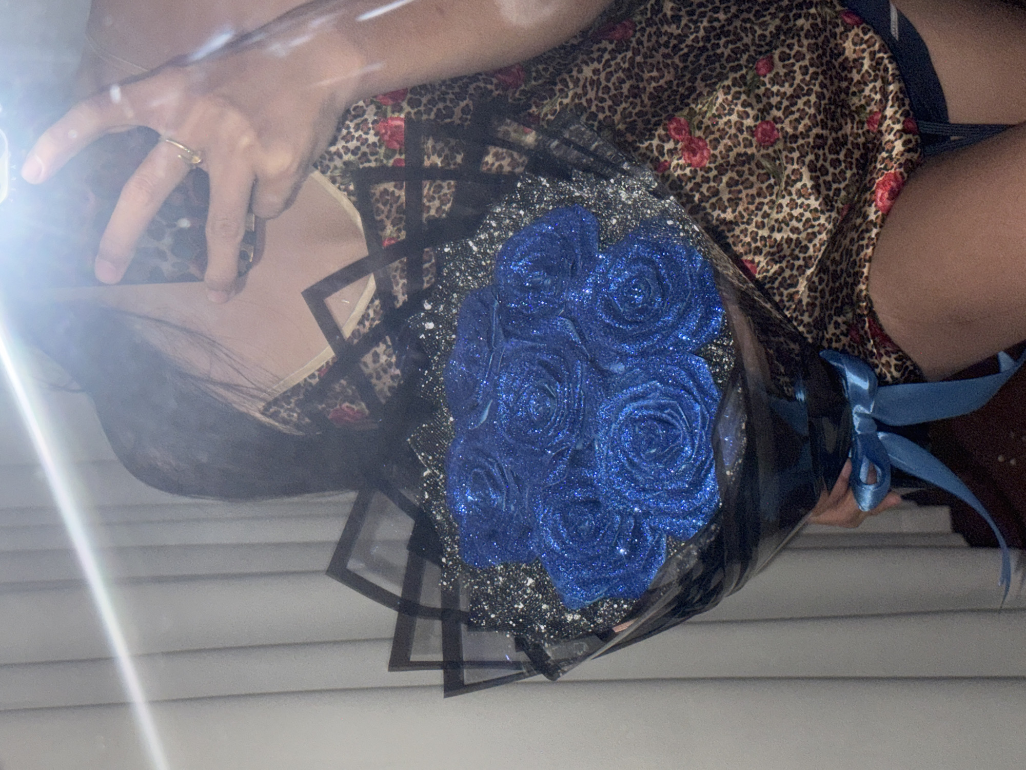 A person is taking a mirror selfie holding a bouquet of blue glitter roses. They are wearing a leopard print slip with red roses. The background appears to be an indoor setting with vertical blinds. The image was taken in Davao City, Philippines.