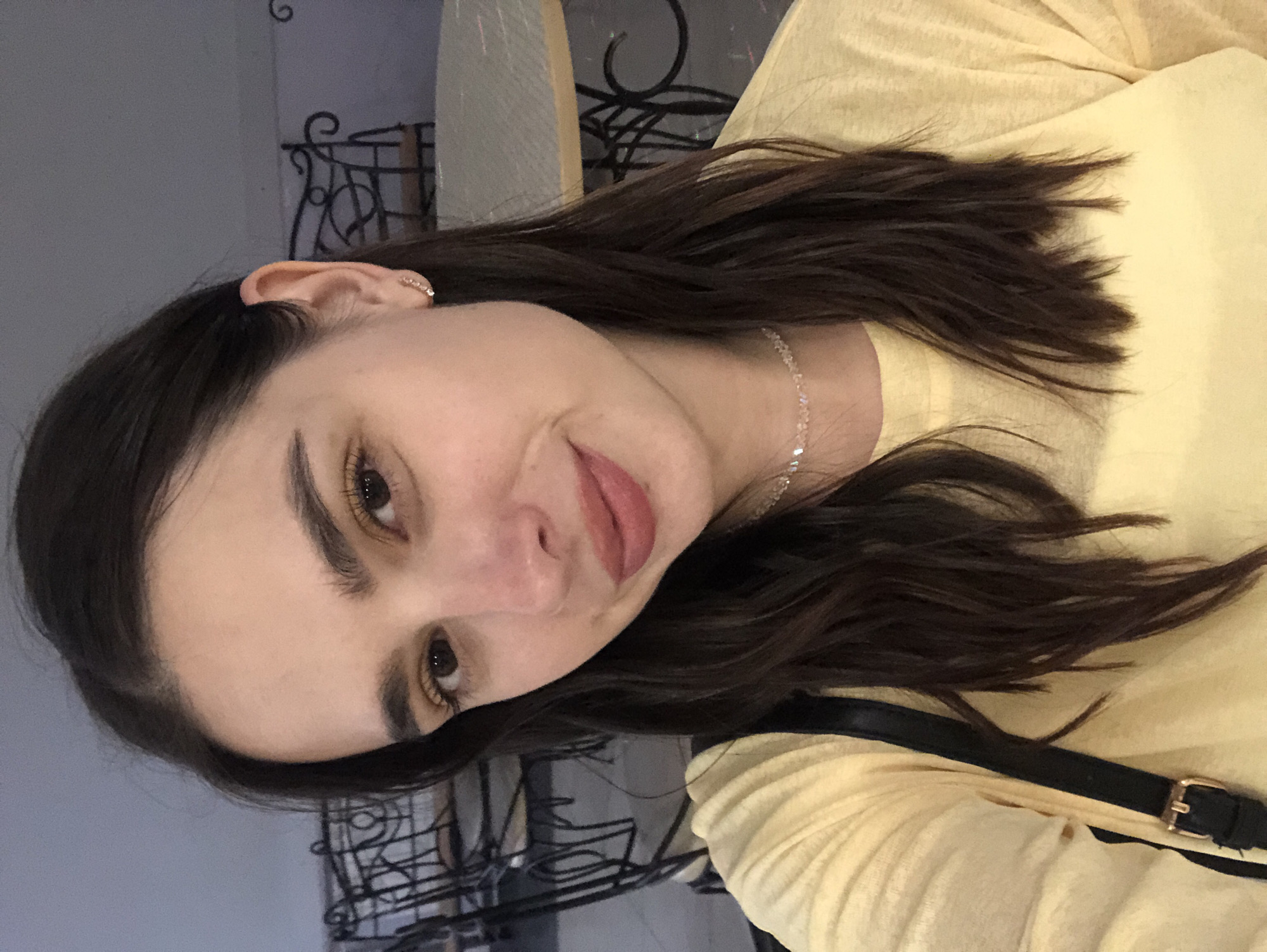A medium close-up, vertically oriented selfie shows a young woman with dark, wavy hair, wearing a light yellow shirt and a delicate, sparkly necklace. Her head is tilted, and her gaze is directed towards the camera with a slight smile. She has prominent eyebrows, dark eyelashes, and rosy lips. A small diamond earring is visible in her left ear. In the background, a blurred, dark metal decorative railing is visible, suggesting an indoor setting, possibly a cafe or restaurant with ornate furnishings. The lighting appears to be artificial, creating a soft ambiance. The woman's expression conveys a sense of casual ease and confidence. There is no visible text in the image. The overall impression is a candid and personal snapshot.