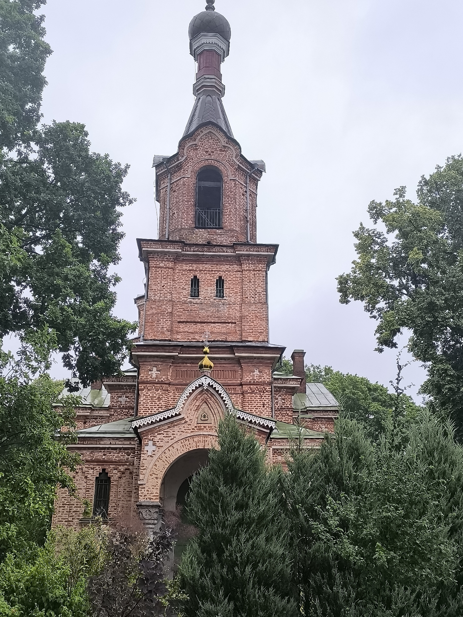 The image shows the exterior of a brick church in Kuldīga, Latvia. The church features a tall, multi-tiered bell tower with a dark dome and a red and grey spire. The facade is adorned with architectural details such as arched windows, decorative brickwork, and a golden dome on a smaller structure above the entrance. The building is partially obscured by lush green trees and foliage, suggesting it is set in a natural or park-like environment. The sky is overcast and pale, indicating a cloudy day. There are no people visible in the image. The overall scene conveys a sense of historical architecture surrounded by nature.