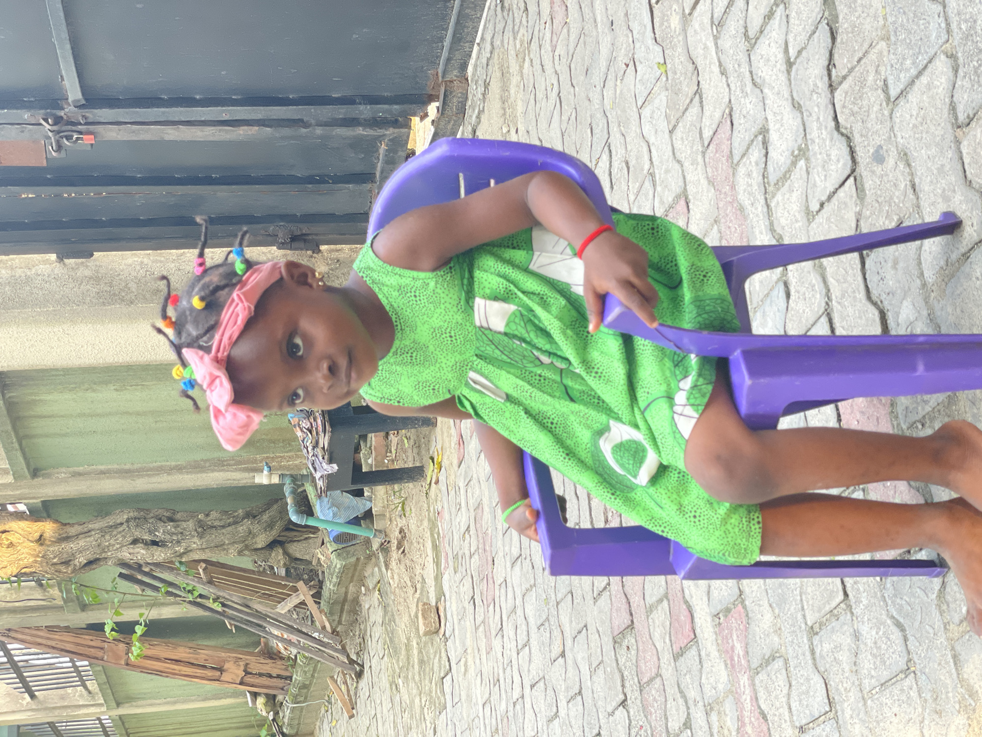 A young girl, approximately 4-6 years old, is seated in a small purple plastic chair on a paved surface in an outdoor residential or semi-urban area in Ado, Nigeria. She wears a sleeveless green dress with a patterned design, a pink headwrap, and colorful plastic beads in her hair, which is styled in multiple short braids. Gold stud earrings are visible in her ears, and she wears a green bracelet on her right wrist and a red string bracelet on her left wrist. Her bare feet rest on the light gray and reddish-brown interlocking paver stones. The girl is looking towards the viewer's upper right with a neutral expression.
To her left (viewer's right), a dark metal gate with a red padlock is partially visible. Behind her, a textured green wall spans the background. Along the wall, to her right (viewer's left), is a large, gnarled tree trunk, surrounded by various discarded wooden planks and debris. A blue pipe fixture and a small dark table with miscellaneous items are also present near the wall. The environment suggests an unkempt outdoor space.