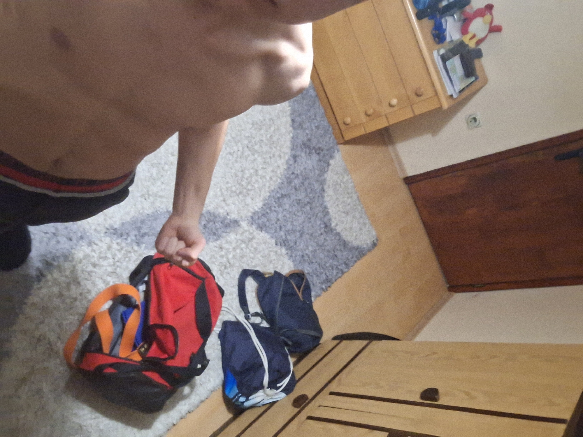 A shirtless individual, wearing black shorts with a red waistband, stands in a room with a light-colored carpet that has a pattern of gray abstract shapes. The person is holding a red and black backpack. Two navy blue bags sit on a wooden surface. A light brown wooden cabinet is attached to the wall, and a wooden door is visible further down the wall. The scene appears to be indoors, potentially in a bedroom or living area. It is not possible to identify the location as Burgas, Bulgaria, without more information.