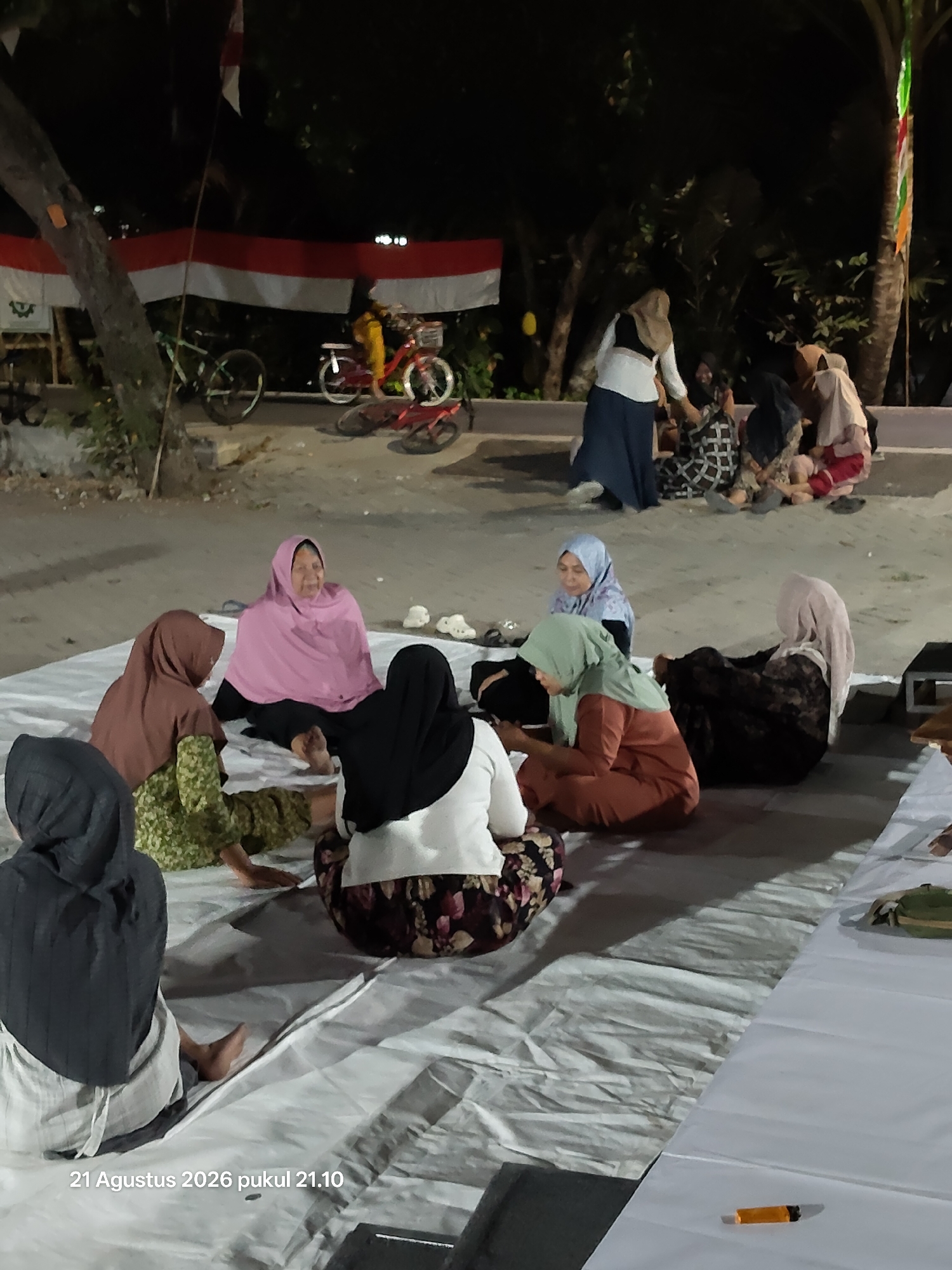 A group of women, some wearing hijabs, are gathered outdoors on a mat at night, in what appears to be the city of Wedi, Indonesia. The Indonesian flag is displayed in the background. Several bicycles are also visible. The women are seated in a circle, some engaged in conversation. The lighting suggests an artificial light source, possibly from a nearby building or portable lights. The overall atmosphere seems relaxed and informal.