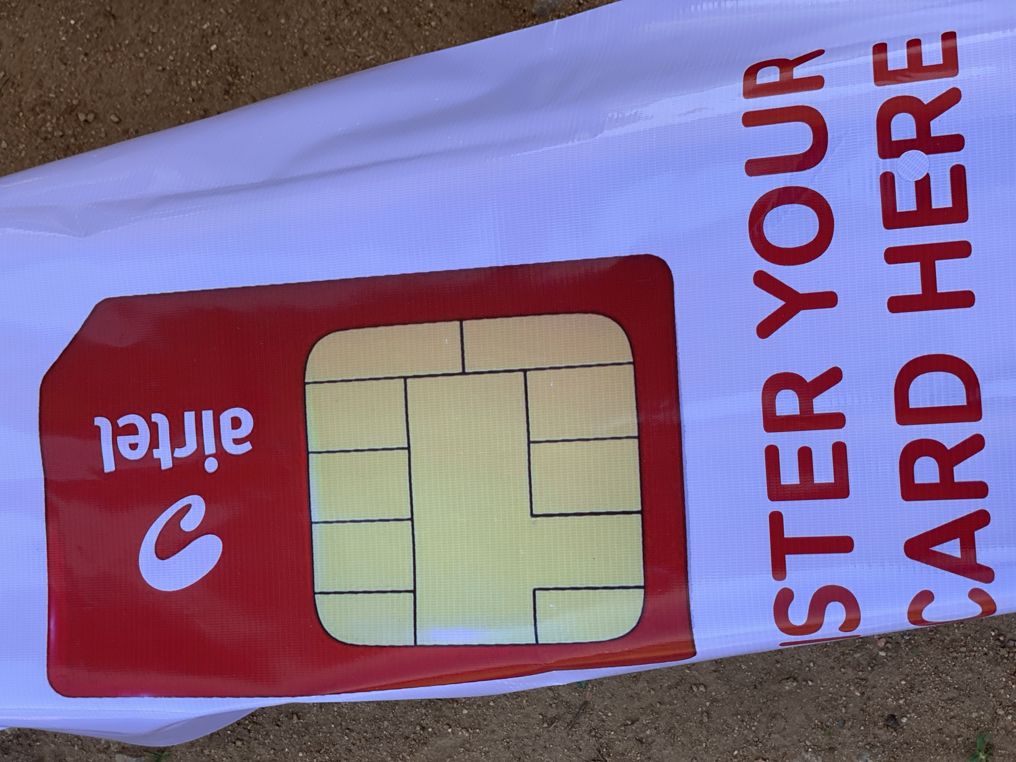 The image shows a close-up, angled view of a large banner advertising Airtel SIM cards in Jalingo, Nigeria. The banner is white and features a large, stylized red SIM card with a yellow chip area outlined in black. The Airtel logo, a white curved shape, is prominently displayed on the red part of the SIM card, next to the word "airtel" in white text.
To the right of the SIM card graphic, red text is printed vertically on the white banner. The text reads "REGISTER YOUR SIM CARD HERE". The letters are bold and appear to be made of vinyl or a similar material. A small, circular hole is visible in the "H" of "HERE," suggesting the banner might have been previously mounted or is designed to be hung.
The banner is laid out on a surface of brown dirt and gravel, with small green plants visible at the bottom edge of the frame. The lighting suggests it is daytime, and the overall scene appears to be outdoors. The focus is entirely on the banner and its advertising message, with no people or other distractions visible.
