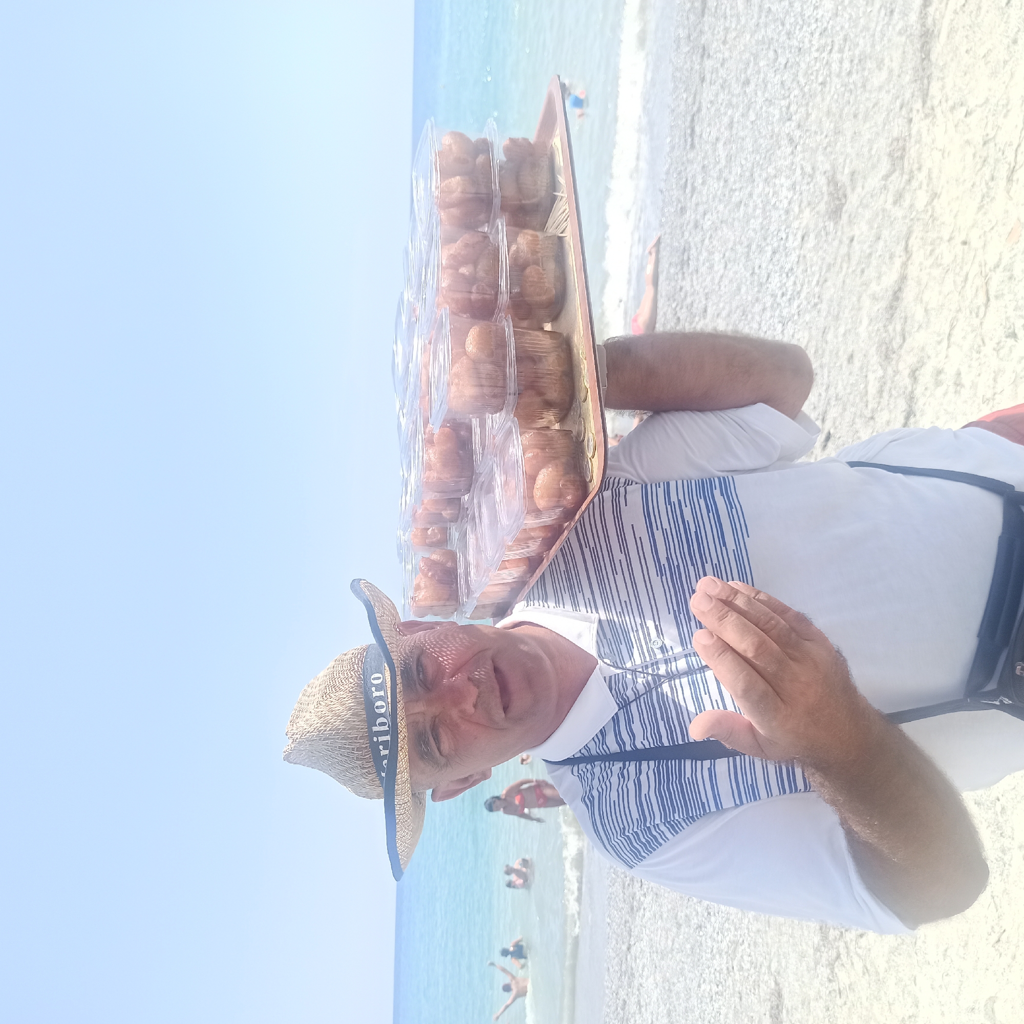 In a beach scene, a man wearing a straw hat with the word "riboro" on its band and a white polo shirt with blue stripes holds a tray of small, round, fried pastries. He is standing on a pebbly beach with the sea and sky in the background, and other beachgoers are visible in the water. The man's left hand is raised, palm open, as if offering the pastries. The overall lighting suggests a sunny day. The image appears to be taken in the vicinity of Ωραιόκαστρο, Greece, known for its coastal areas.