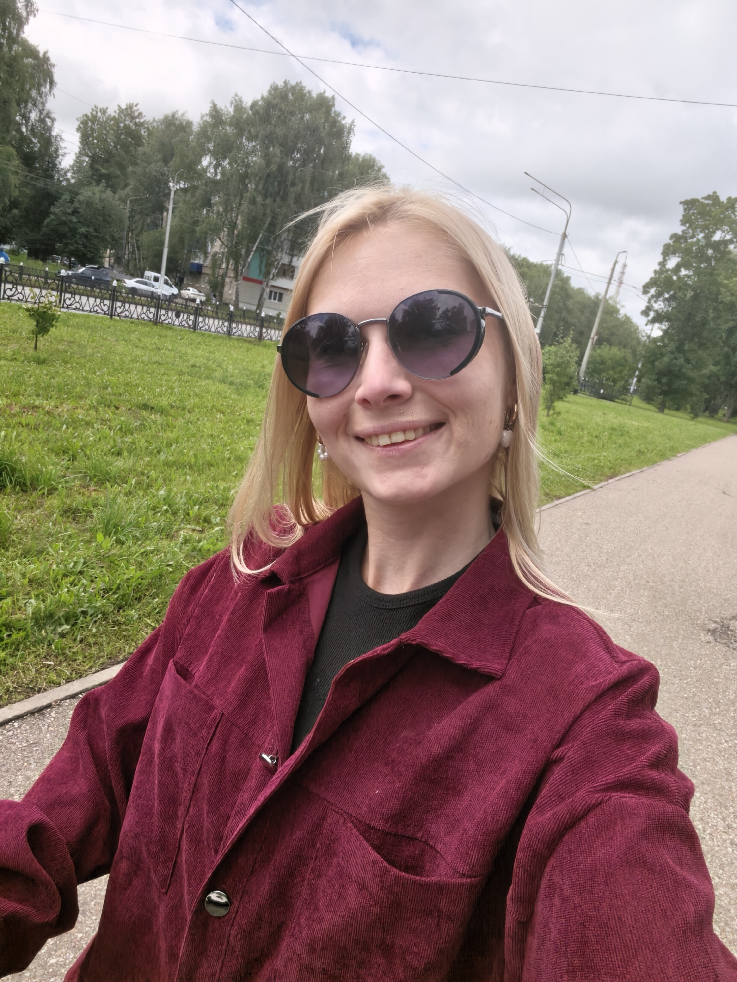 A young woman with blonde hair and sunglasses takes a selfie outdoors in Sterlitamak, Russia. She is wearing a dark red corduroy jacket over a black top and a pearl earring on her right ear. She is smiling and looking at the camera. The background shows a grassy park area with trees and a paved path. There are some cars and buildings visible in the distance. The sky is cloudy.