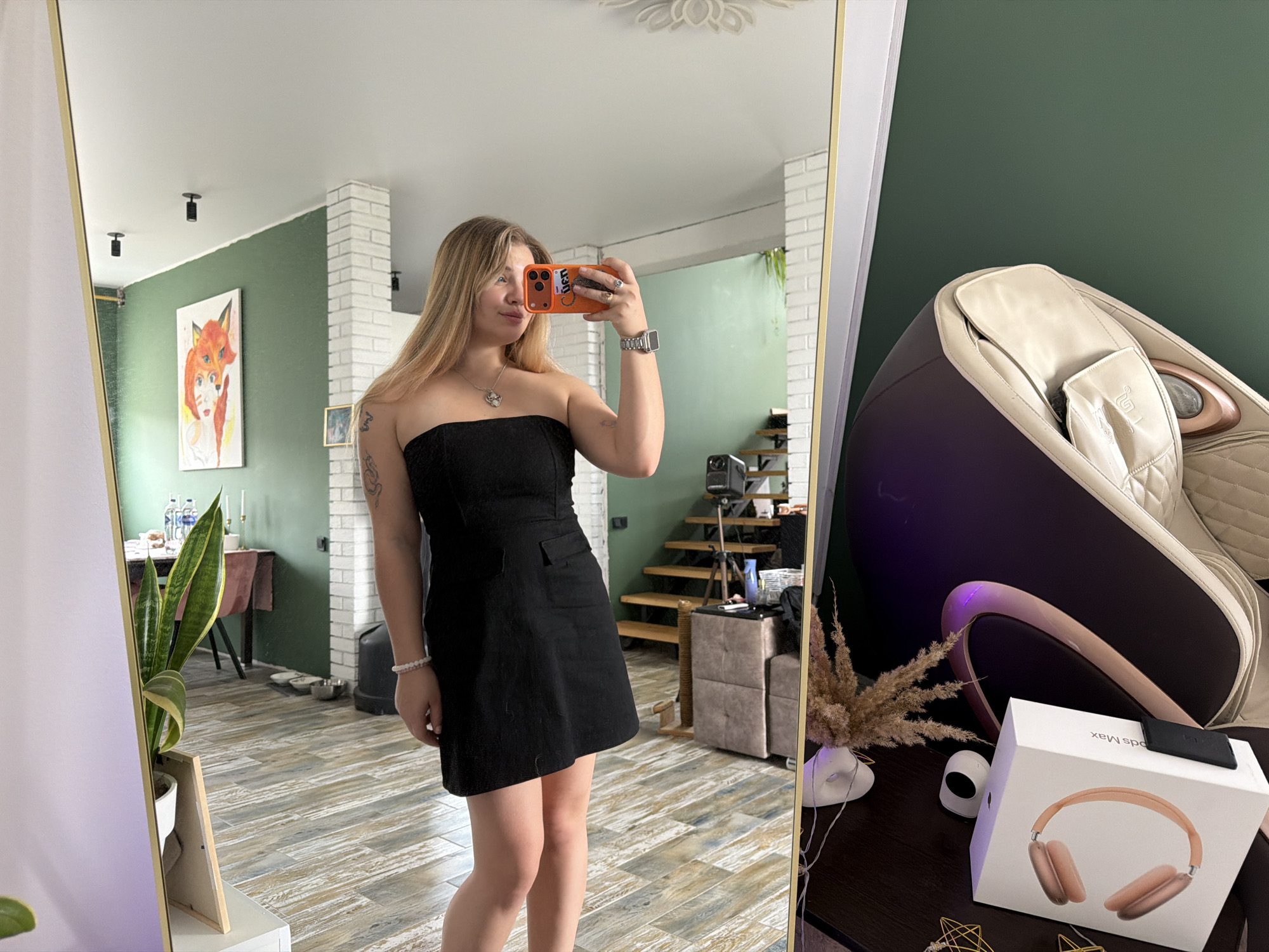 A waist-up view shows a woman in a black dress taking a selfie in a mirror. Her phone is orange, and she wears a silver wristwatch. The reflection reveals a room with wooden floors, a white brick-like wall, and green walls. In the background are a dining table and a staircase. A painting of a fox adorns the wall. Next to the mirror is a massage chair, a box of headphones, a vase with dried flowers, and a smart home speaker. The setting is a room in a building in Большая Шильна, Russia.