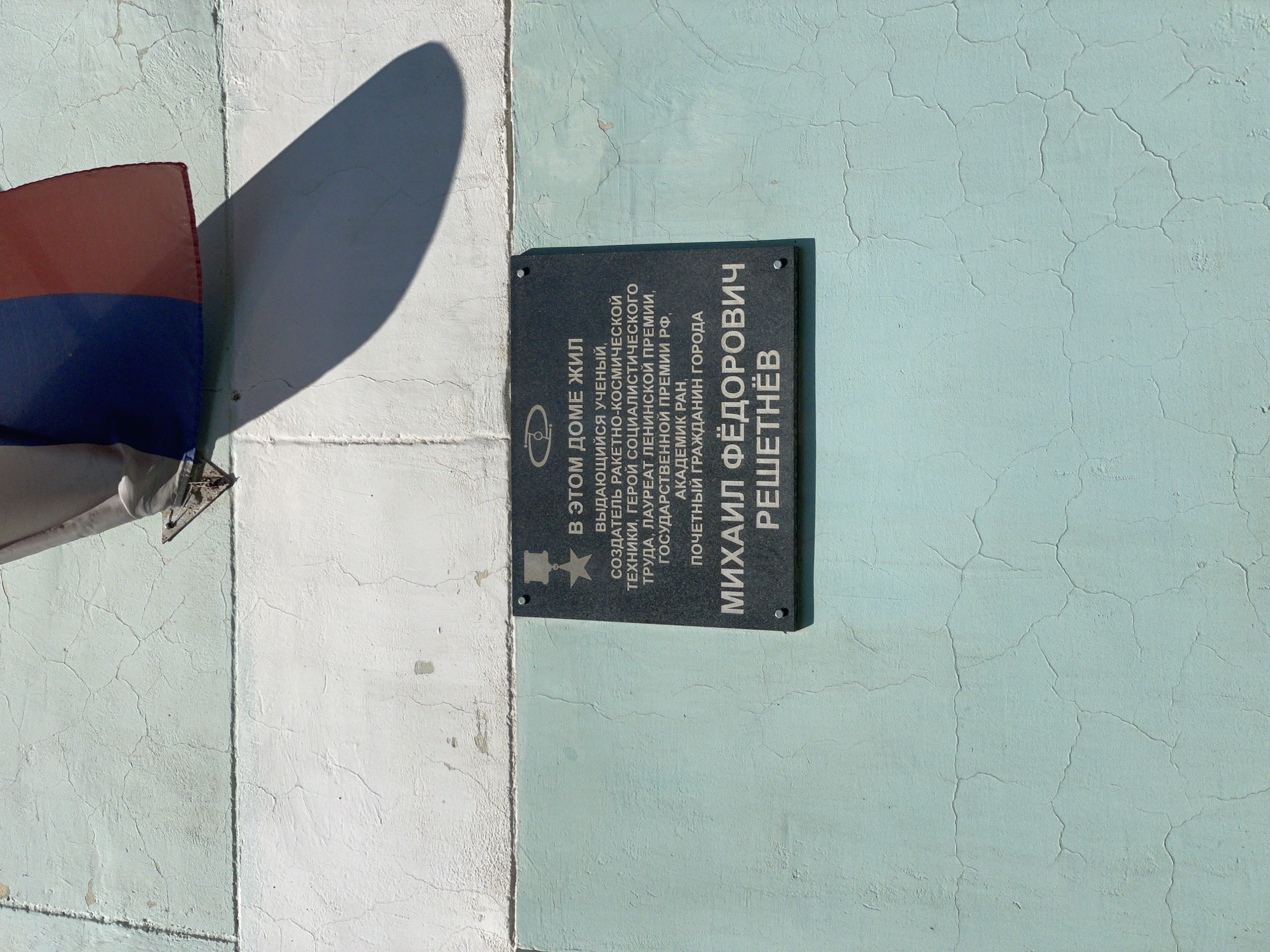 The image shows a memorial plaque on a cracked, pale green and white wall. A Russian flag is partially visible at the top, casting a shadow. The plaque is dark gray and mounted with four screws. The text on the plaque is in Russian and reads: "В ЭТОМ ДОМЕ ЖИЛ ВЫДАЮЩИЙСЯ УЧЁНЫЙ, СОЗДАТЕЛЬ РАКЕТНО-КОСМИЧЕСКОЙ ТЕХНИКИ, ГЕРОЙ СОЦИАЛИСТИЧЕСКОГО ТРУДА, ЛАУРЕАТ ЛЕНИНСКОЙ ПРЕМИИ, ГОСУДАРСТВЕННОЙ ПРЕМИИ РФ, АКАДЕМИК РАН, ПОЧЕТНЫЙ ГРАЖДАНИН ГОРОДА МИХАИЛ ФЁДОРОВИЧ РЕШЕТНЁВ". This translates to: "IN THIS HOUSE LIVED AN OUTSTANDING SCIENTIST, CREATOR OF ROCKET AND SPACE TECHNOLOGY, HERO OF SOCIALIST LABOR, LAUREATE OF THE LENIN PRIZE, STATE PRIZE OF THE RUSSIAN FEDERATION, ACADEMICIAN OF THE RUSSIAN ACADEMY OF SCIENCES, HONORARY CITIZEN OF THE CITY MIKHAIL FEDOROVICH RESHETNEV." The plaque is dedicated to Mikhail Fedorovich Reshetnev, a prominent figure in Soviet and Russian space technology and a former honorary citizen of Zheleznogorsk, Russia. The wall's condition suggests age and exposure to the elements.