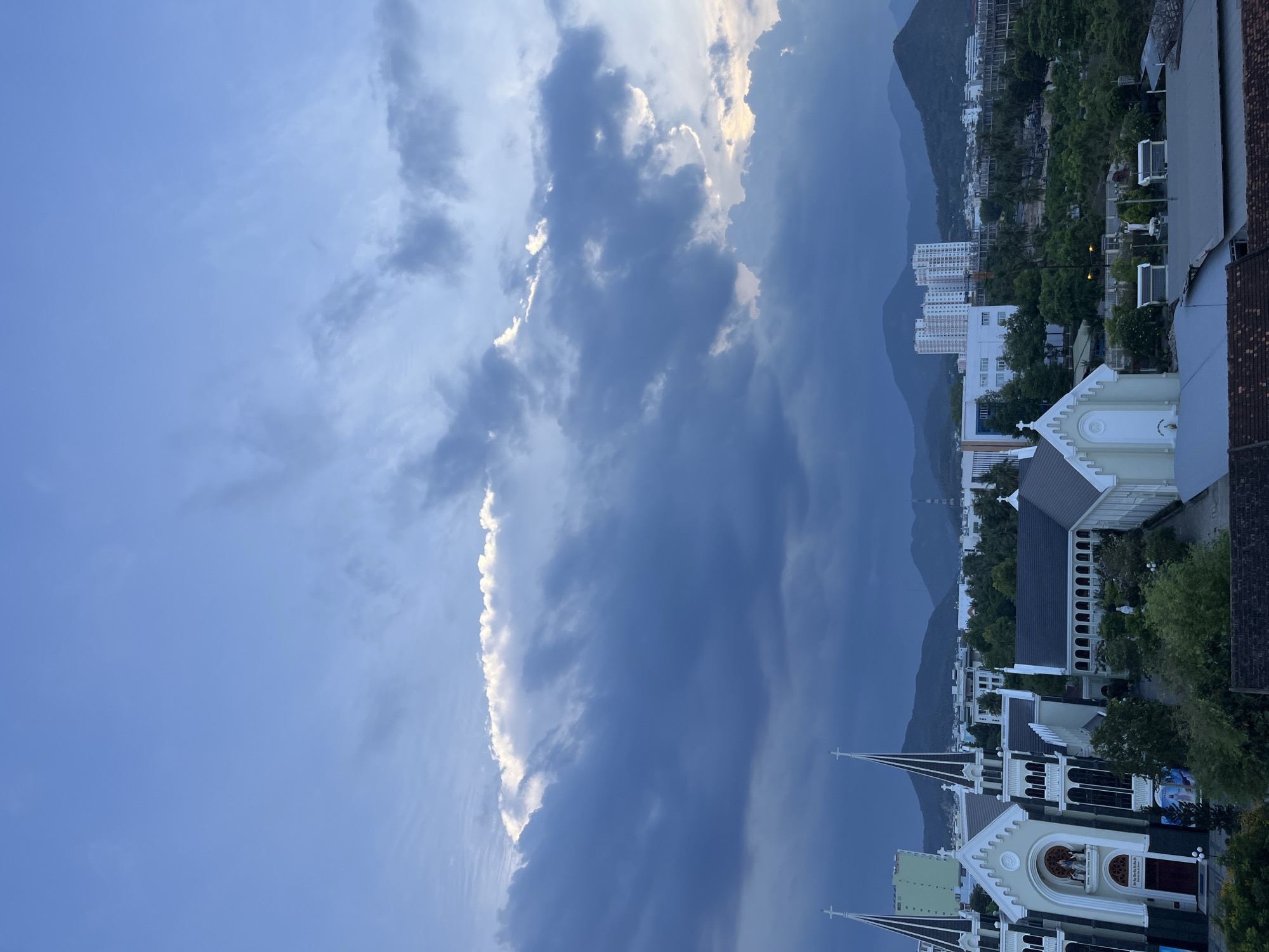 The image shows a cityscape in Bac Nha Trang Ward, Vietnam, taken from an elevated position. The sky is mostly blue with significant cloud cover, including a distinct strip of bright white clouds. In the middle ground are mountains. In the foreground are church buildings with white trim and spires. Beyond the church are various buildings and vegetation, indicating a populated area.