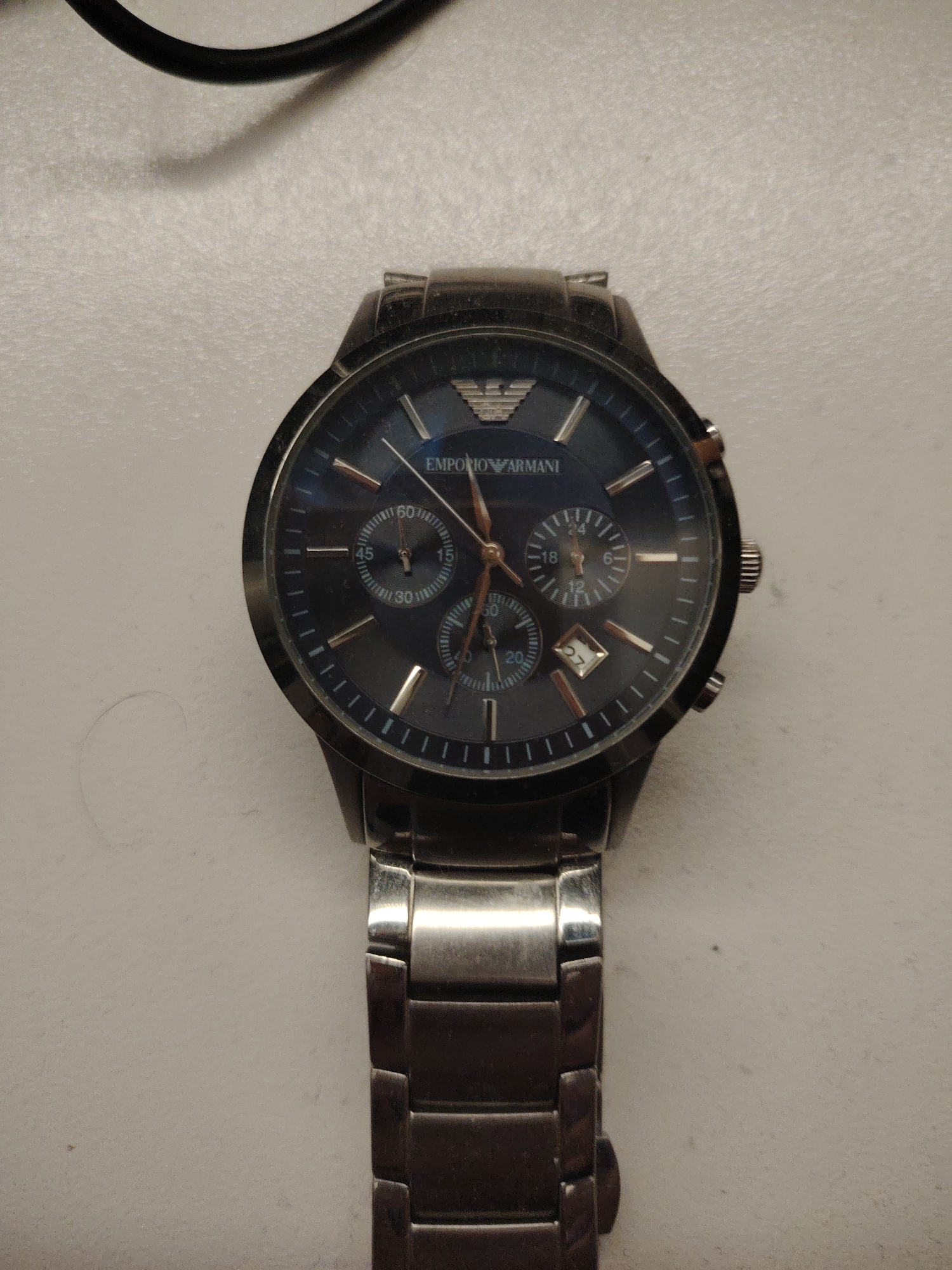 A metallic chronograph wristwatch, identified as an Emporio Armani, is centrally positioned on a light, off-white, textured surface. The watch features a dark, likely gunmetal-toned, circular case and a matching three-link metallic bracelet.
The deep blue-grey dial exhibits a subtle sunburst finish. It is marked with applied metallic baton hour markers, with double markers at the 12 o'clock position. The hour, minute, and sub-dial hands are metallic, appearing rose gold or copper-toned. Three sub-dials are present: a large 60-unit counter at 9 o'clock (marked 15, 30, 45, 60), a 24-hour or small seconds counter at 3 o'clock (marked 6, 12, 18, 24), and a smaller sub-dial at 6 o'clock (marked 20, 40, 60). A date aperture at the 4:30 position displays the number '27' against a white background. The watch's right side shows a knurled crown flanked by two pushers for the chronograph function. Minor scuffs and wear are visible on the watch case and bracelet links.
The light-colored background surface, possibly a textile or paper, exhibits minor surface imperfections and dust specks. In the upper left corner of the frame, a dark, curved cable or wire is partially visible, casting a faint shadow on the surface. No people or specific actions are depicted. The environment provides no identifying details for a specific city or region.