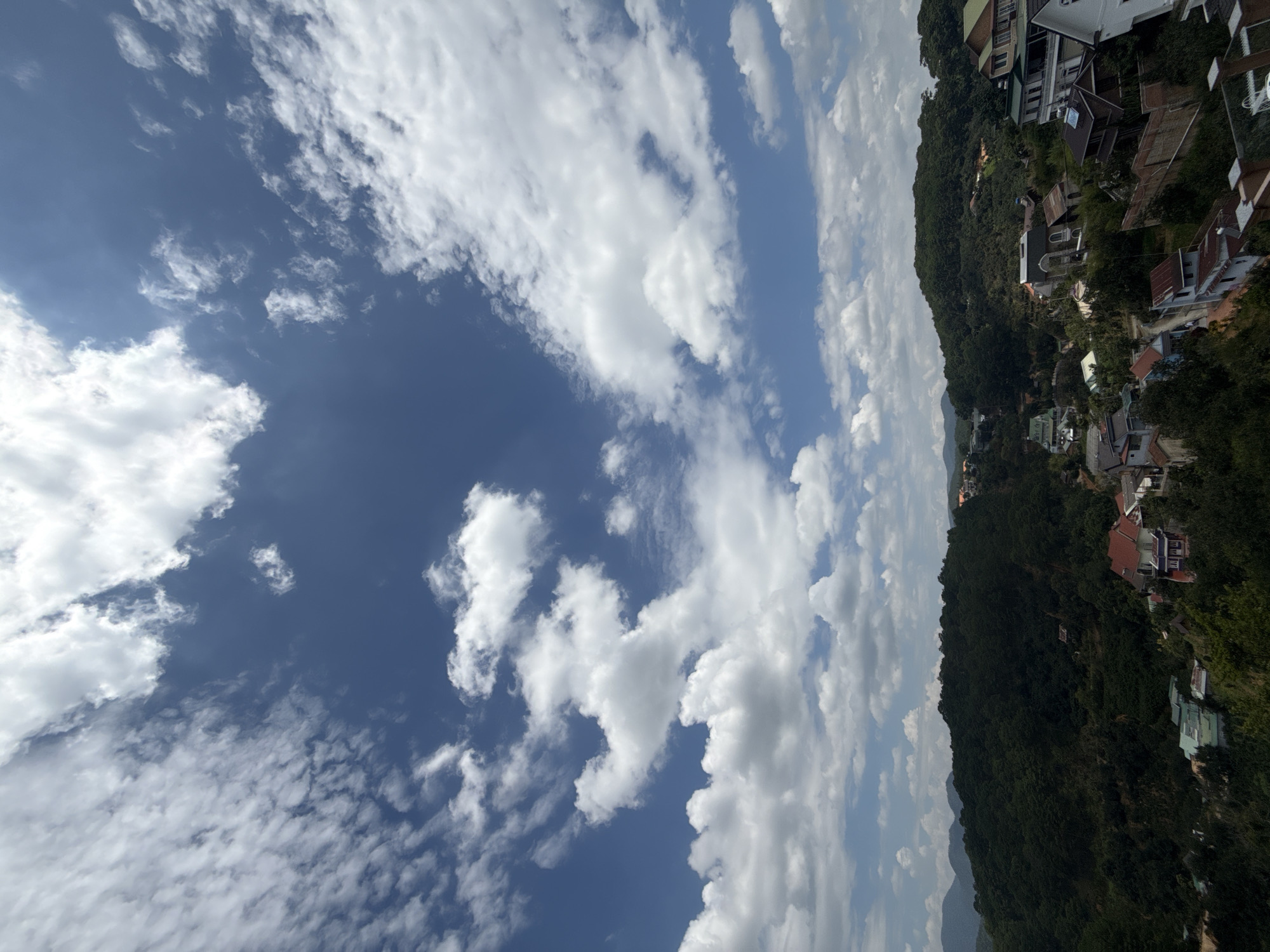 The image depicts a panoramic view of the city of Da Lat, Vietnam, characterized by its hilly terrain and lush vegetation. In the foreground, several houses are nestled amidst dense green foliage, some with red tile roofs and others with a more modern architectural style featuring multiple stories and balconies. These houses are scattered across the slopes of the hills, suggesting a residential area built on uneven ground. The midground is dominated by rolling hills covered in thick, dark green trees, giving the impression of a densely forested landscape. In the distance, layers of hazy, blue-green mountains fade into the horizon, indicating the broader geographical setting of Da Lat. The sky above is a vibrant blue, punctuated by scattered white clouds of varying shapes and sizes. Some clouds are wispy and thin, while others are more voluminous and billowy, creating a dynamic and expansive sky. The lighting suggests it is daytime, likely mid-morning or afternoon, with the sun casting light on the clouds and illuminating the landscape. The overall scene conveys a sense of a tranquil, picturesque city with a strong natural element.