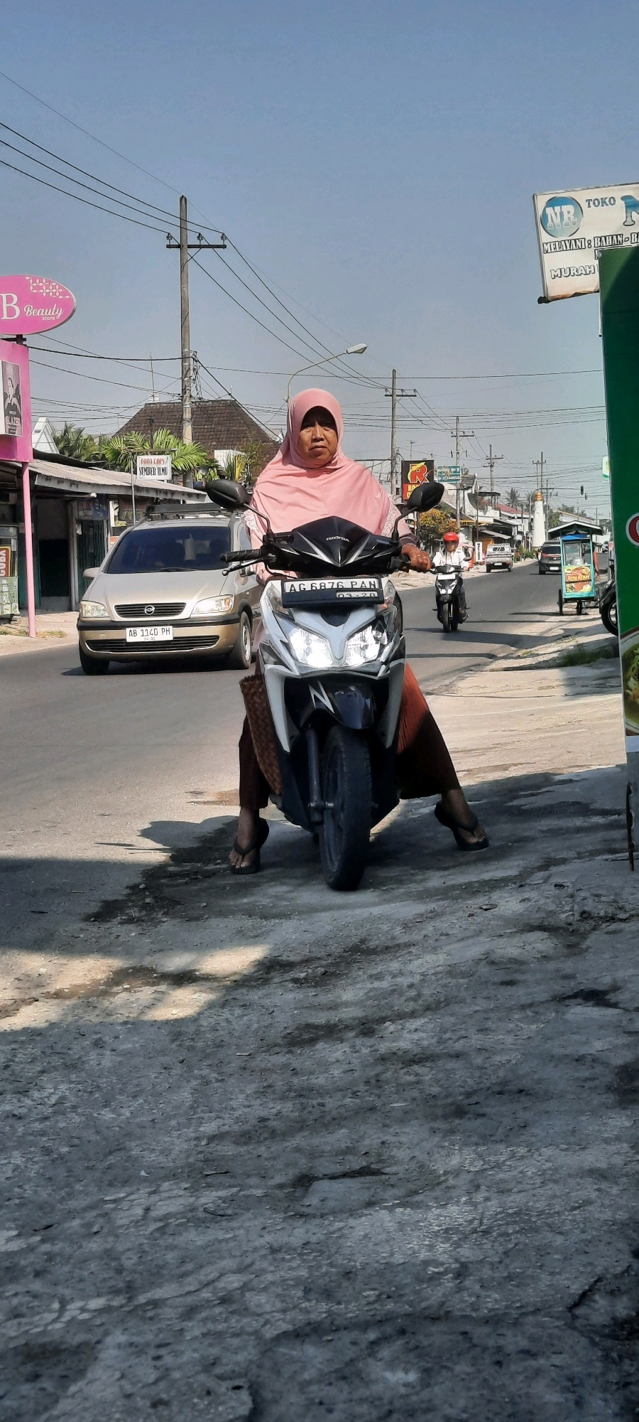 A woman wearing a pink hijab and an orange skirt is sitting on a white and black motorcycle. Her feet are flat on the ground, wearing flip-flops. The motorcycle has a license plate that reads "AG 6876 PAN". In the background, a silver car with license plate "AB 1140 PH" is driving on the road. There are shops and buildings lining the street. Power lines are visible above. The sky is clear and blue. The scene is set in Selopuro, Indonesia.