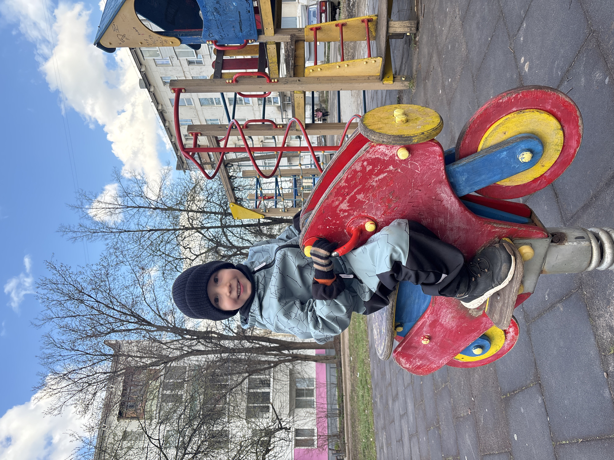 A young child, appearing to be a boy, sits on a brightly colored spring rider on an outdoor playground in Chaikovsky, Russia. The child is dressed in a black beanie, a light blue and grey patterned jacket, dark pants, and black shoes with white soles, along with striped gloves. He is smiling broadly and looking directly at the camera, suggesting enjoyment.
The spring rider is shaped like a motorcycle or scooter, painted red and blue with yellow circular elements for wheels and handles. Its paint is worn in places, indicating frequent use. The ground is paved with grey interlocking bricks.
In the background, a multi-level playground structure made of wood and metal, featuring red and yellow components, is visible. Beyond the playground, bare trees stand against the sky, suggesting late winter or early spring. Several multi-story residential buildings, characteristic of Soviet-era architecture, line the horizon; one distinctively features a pink base. The sky is a clear blue with scattered white clouds, indicating daytime. The scene captures a moment of play on a community playground.
