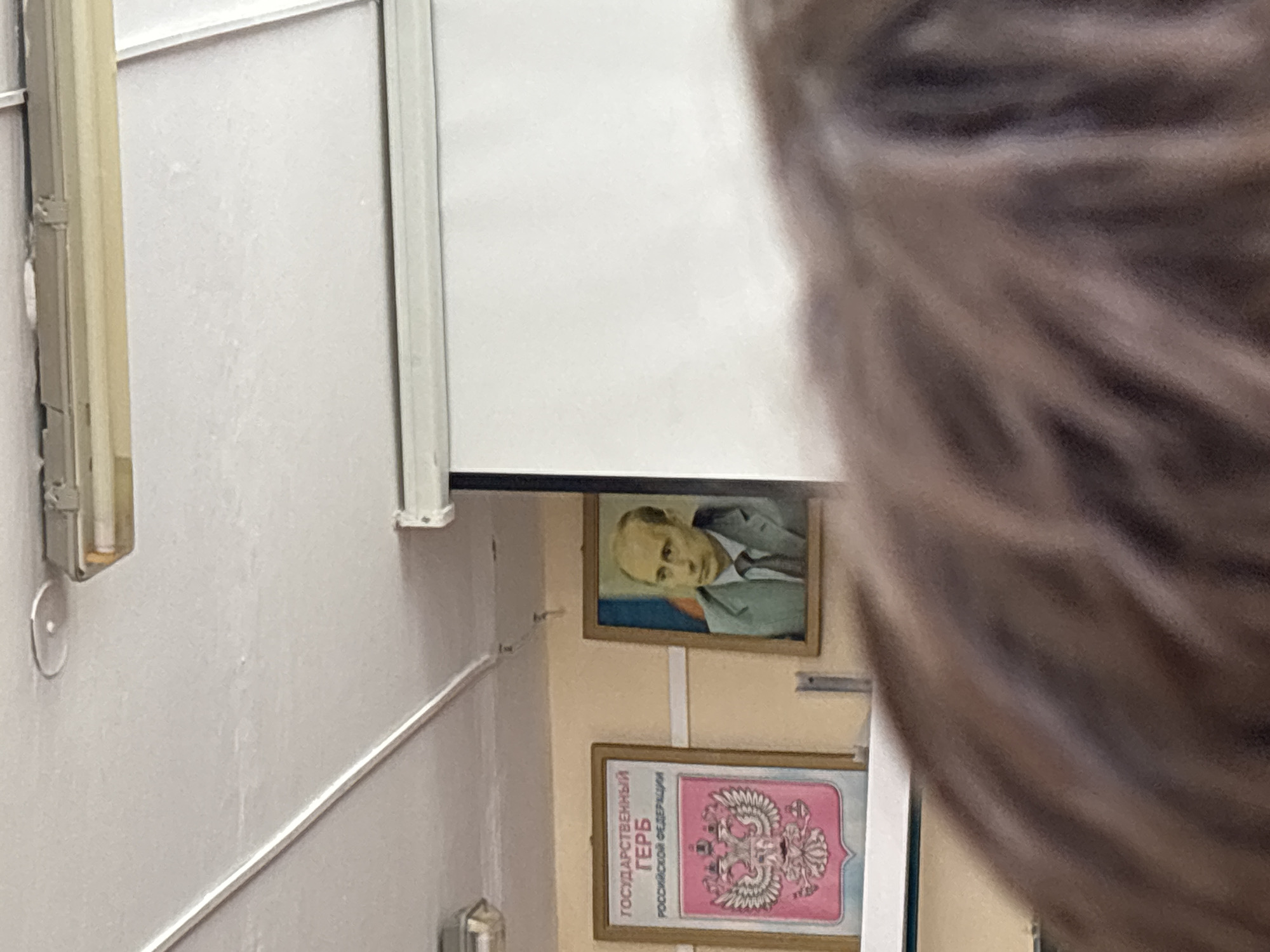A partially obstructed view reveals a wall inside a building in the city of Unknown, Unknown, featuring two framed items. The top frame contains a portrait of a man. Below it, the second frame displays a graphic with text in Russian, including the words "Государственный Герб Российской Федерации" (State Emblem of the Russian Federation). A portion of a person's head is also visible, obscuring part of the scene. The wall is painted in a light color. A metallic fixture is attached to the wall, with a light fixture above.