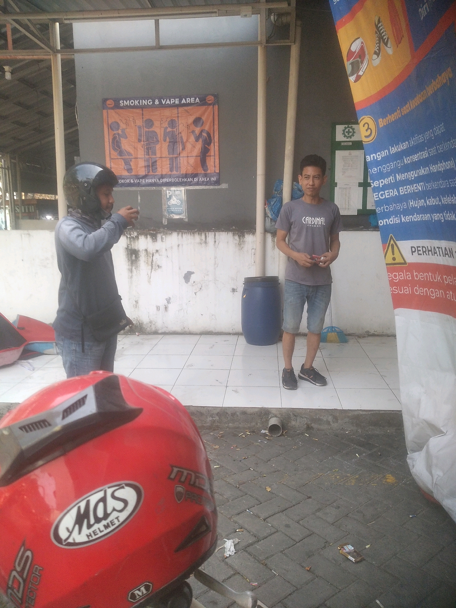 In Cilegon, Indonesia, two men are present in an outdoor smoking and vape area. The man on the left, wearing a helmet and a grey long-sleeved shirt over blue jeans, is holding a clear plastic cup. The man on the right, wearing a grey t-shirt with "CARDINAL" printed on it and denim shorts, is standing with a pack of cigarettes in his hand. Behind them, a blue barrel and a broom are visible, along with a "SMOKING & VAPE AREA" sign depicting people smoking. A large banner with Indonesian text and safety warnings, including a triangle warning symbol, is positioned on the right side of the frame. The ground is paved with grey interlocking bricks, and cigarette butts are scattered across it. In the foreground, a red motorcycle helmet with "Mds HELMET" branding is prominently displayed. The environment suggests a public area, possibly a rest stop or a designated zone within a facility.