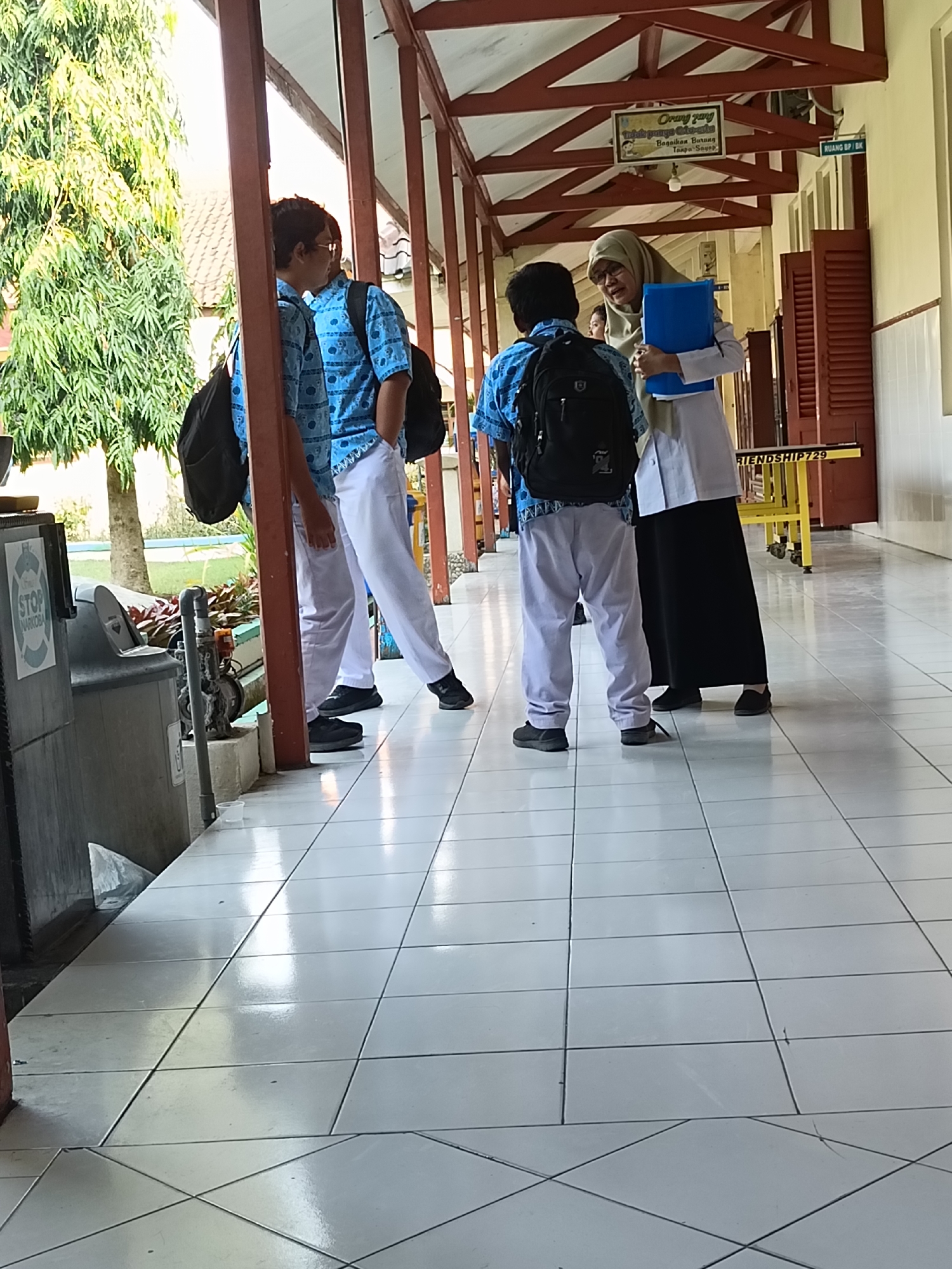 The image depicts a hallway of a school in Kediri City, Indonesia, likely in the afternoon due to the bright sunlight. In the foreground, two male students wearing blue patterned shirts and white pants with black backpacks are standing. One student is looking away from the camera, while the other faces a female teacher. The teacher, wearing a hijab and a light-colored uniform, is holding a blue folder and appears to be speaking to the student. Her black skirt and shoes are visible. The hallway has a tiled floor and is supported by red wooden pillars. In the background, a yellow cart with wheels is visible, along with a sign that reads "RUANG BP / BK." There is also a sign on the wall with text in Indonesian, including "Orang Yang Berakhlaq Mulia," and another smaller sign with the word "STOP NARKOBA." A large tree with green foliage is visible outside the hallway to the left.
