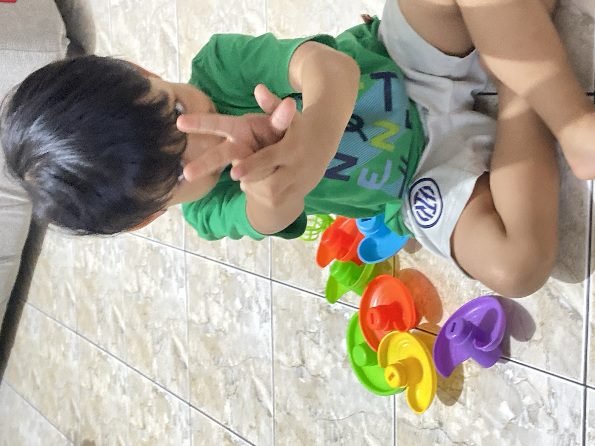A young boy wearing a green t-shirt and light-colored shorts is sitting on a tiled floor. He is holding up two fingers in a peace sign towards the camera. In front of him, there are several colorful plastic toys, including what appear to be parts of a water toy, in shades of yellow, orange, red, green, blue, and purple. The boy's face is partially obscured by his hands and hair. The environment appears to be indoors, with light-colored tiled flooring. This image was taken in Margorejo, Indonesia.
