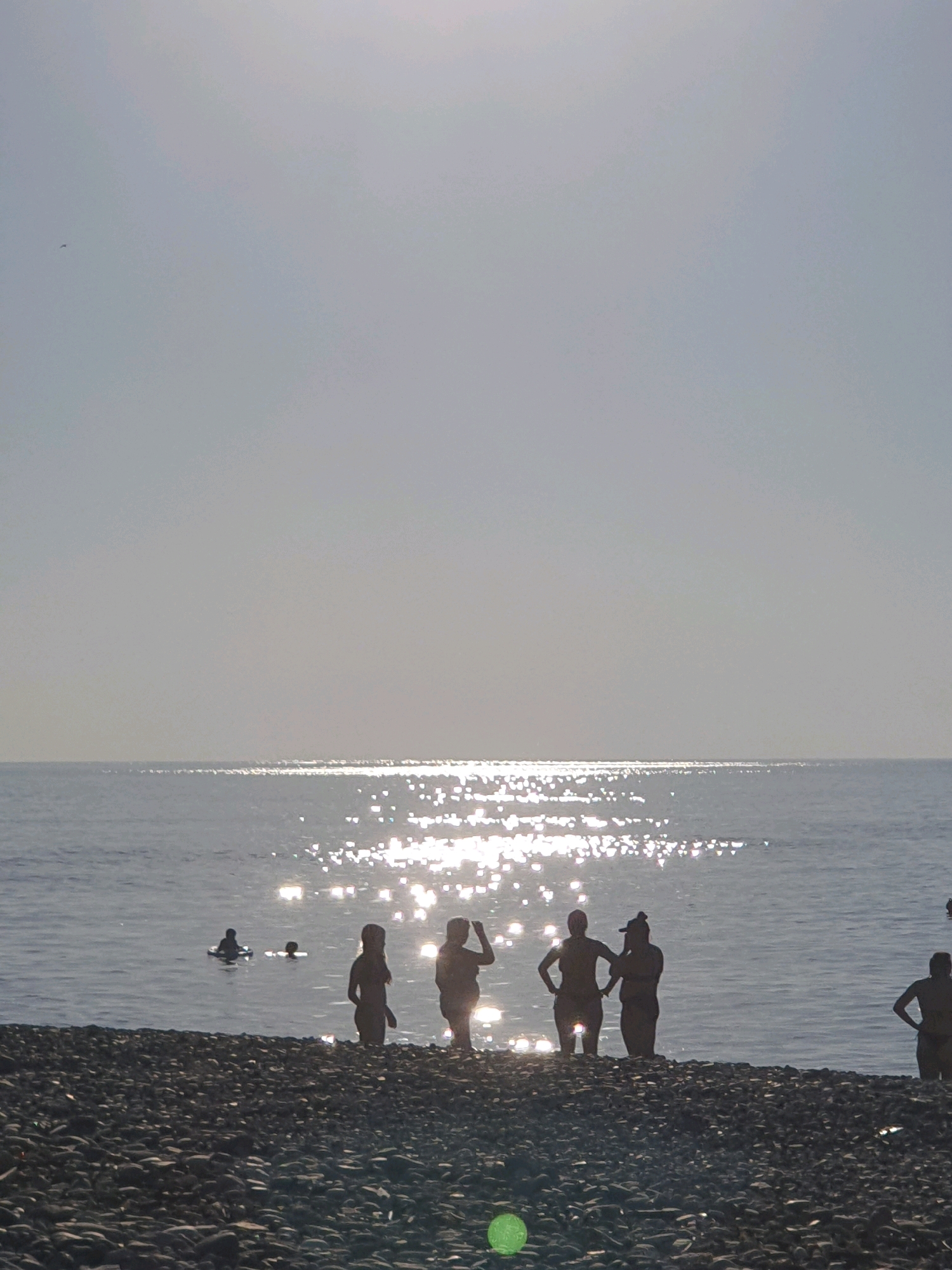 The image depicts a silhouette of several people on a pebble beach with the Black Sea in the background. The sun is shining brightly, creating a shimmering reflection on the water. In the foreground, a dense arrangement of dark pebbles covers the beach. Several individuals are visible standing in the shallow water or on the edge of the water. Among them, a group of four appears to be posing or interacting. Further out in the water, two individuals are seen in what appears to be swimming or playing. The sky is bright and somewhat hazy, indicating a sunny day. The scene suggests a typical summer day at a beach resort. Based on the characteristic pebble beaches and the presence of palm trees, this location is consistent with the coastline of Batumi, Georgia.