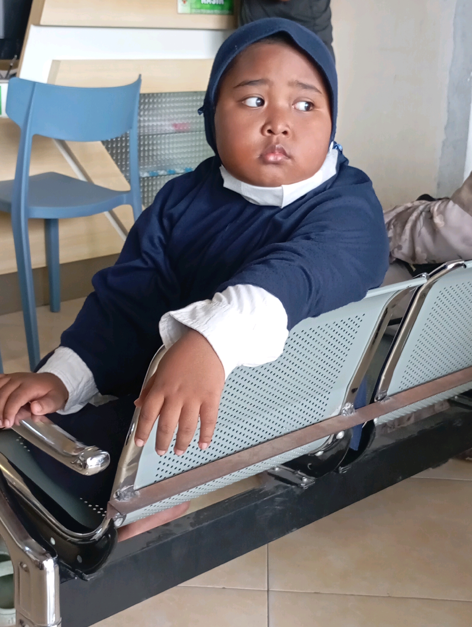 A young child with a round face and wearing a dark blue hijab and white mask is seated on a metal and plastic bench. The child's body is angled to the left, with their head turned to look to the right. Their right arm is resting on the bench, and their left arm is bent at the elbow with their hand near their chest. The bench is situated in an indoor waiting area, with a light blue chair visible in the background. The environment appears to be a public space, possibly a clinic or government office, given the seating arrangement and general decor. The floor is tiled with beige squares. The child's skin tone is dark brown. The overall lighting suggests an indoor setting with artificial light sources.