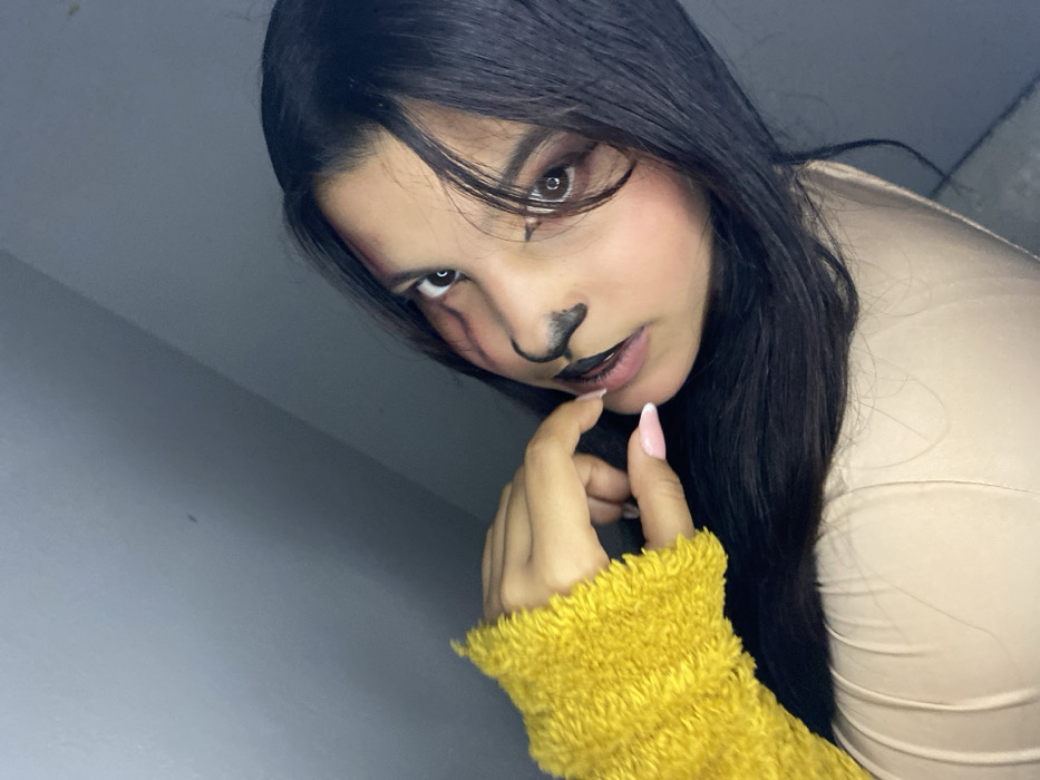A dark-haired woman poses with animalistic makeup, including a black nose and markings around her eyes. She wears a yellow, fuzzy sleeve and light-colored top. She touches her lip with her finger and looks at the camera. The background is a simple, light-colored wall. The lighting is soft, casting shadows that enhance the depth of the shot.