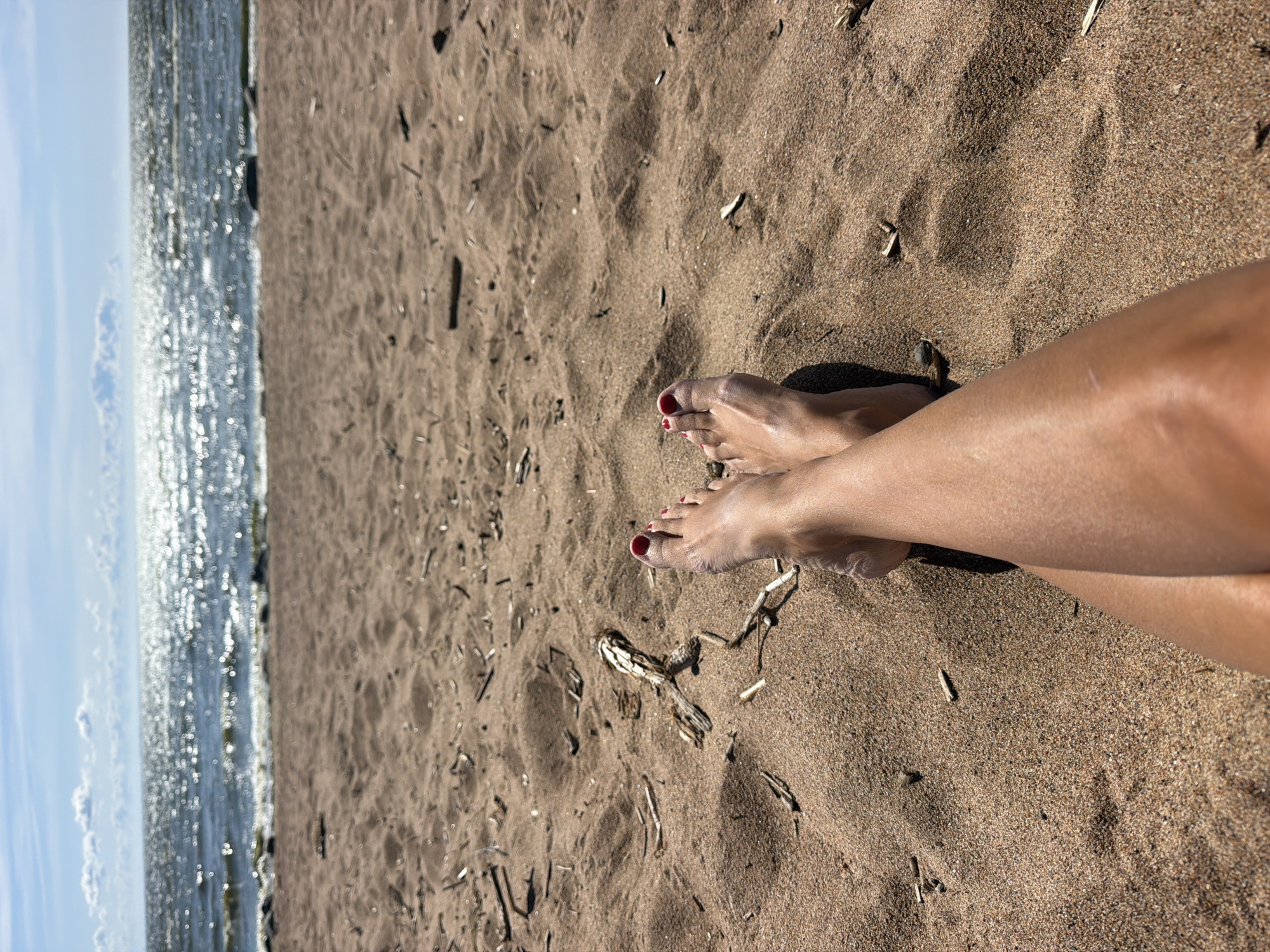A low-angle, medium shot captures a person's legs and feet resting on a sandy beach, with the Gulf of Finland in the background, located in Repino, Russia. The sand is a light brown, with scattered small pieces of driftwood and debris. The person's feet are positioned in the foreground, with toes painted red, crossed over each other. Sunlight glints off the water, which is a deep blue and appears choppy. The sky is a pale blue with scattered white clouds.