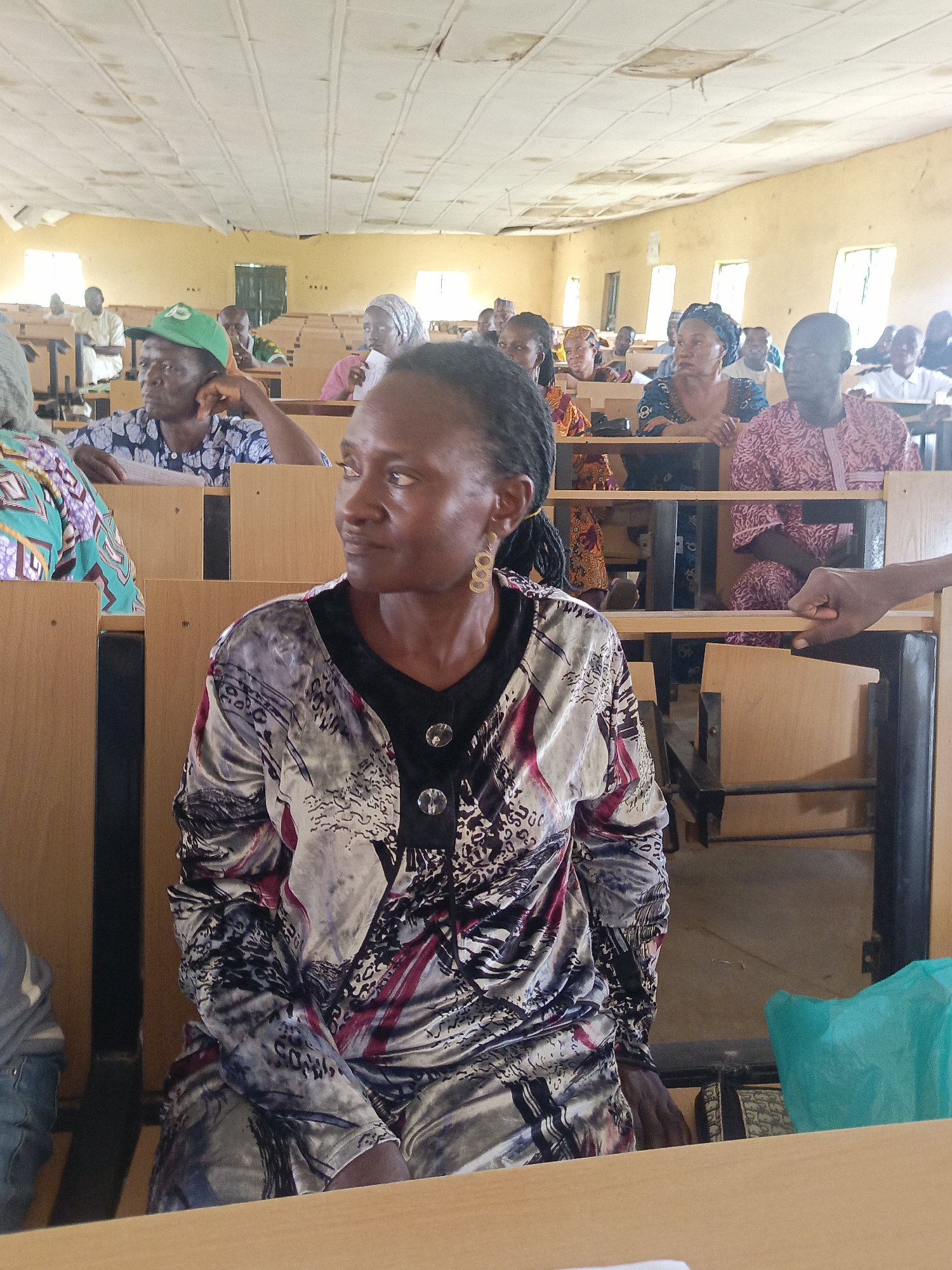 A classroom in Akwanga, Nigeria, is filled with people seated at wooden desks. A woman in the foreground wears a patterned, long-sleeved shirt and gold earrings. Behind her, rows of people are also seated at desks, some wearing colorful traditional clothing and head coverings. The classroom appears to be in a state of disrepair, with a damaged ceiling and aged walls. Light comes in from windows.
