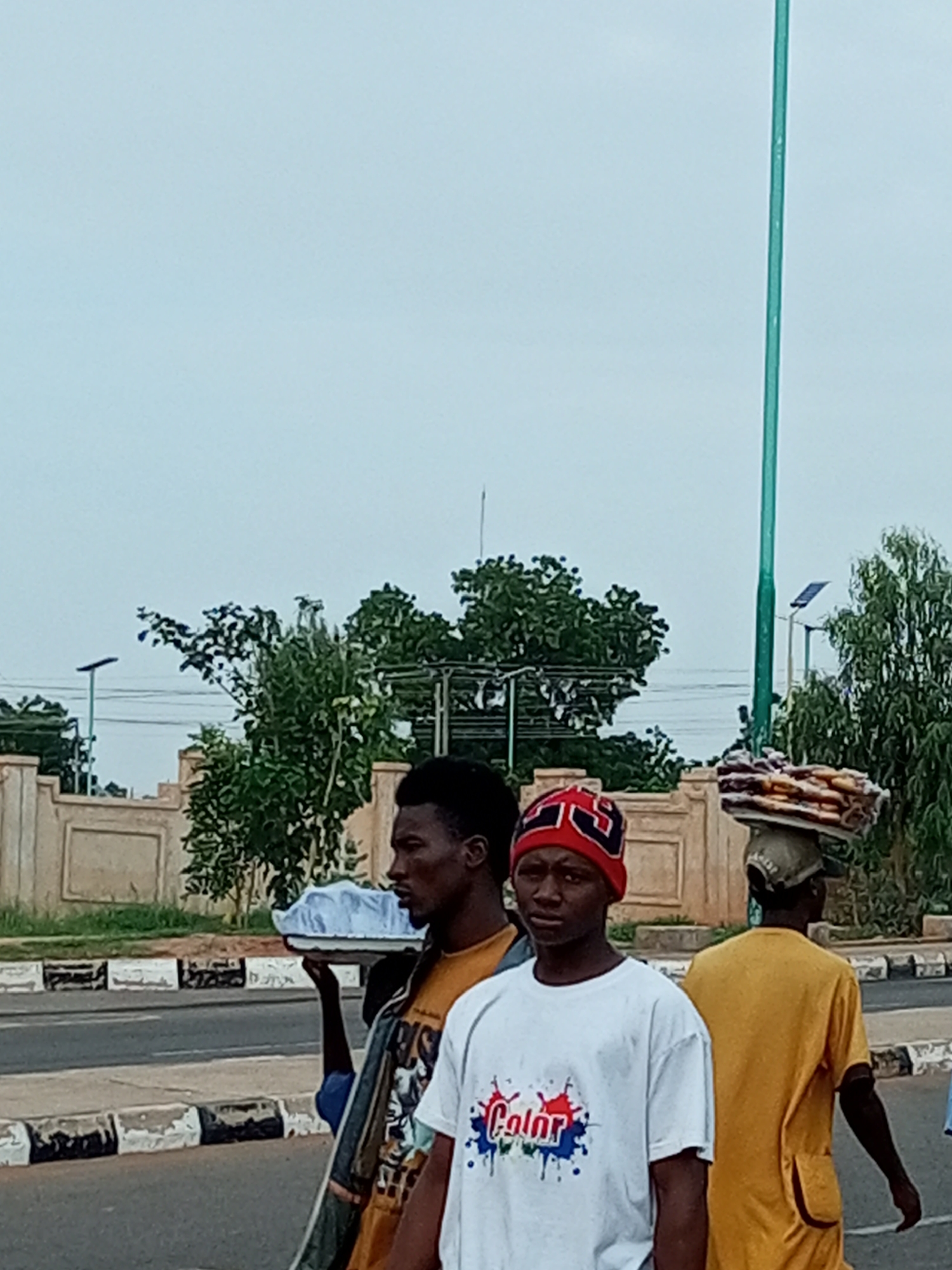In the city of Unknown, Unknown, three young men are seen on a sidewalk next to a road. The man in the foreground on the left, wearing a red and black beanie, has dark skin and is looking towards the camera. He is wearing a white t-shirt with a colorful "Color" design. To his right, a man with dark skin, wearing a yellow t-shirt and a green and white patterned shirt underneath, is walking away from the camera. He is carrying a white tray with a light blue object on it. Behind him, another man, also with dark skin, is walking away from the camera and carrying a stack of items on his head. The background shows a beige wall with decorative panels, green trees, and a tall green pole. The sky is overcast.