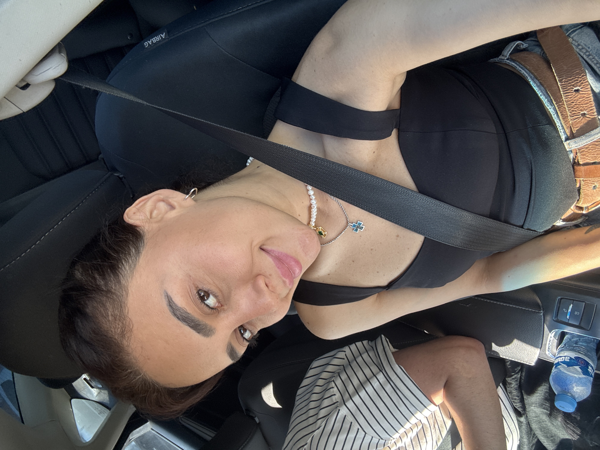 In a car, a woman with dark hair and brown eyes is taking a selfie. She is wearing a black tank top, blue jeans, and two brown belts. A pearl necklace with a cross pendant is visible around her neck. A seatbelt is securely fastened across her chest. Next to her, a person in a striped shirt is visible. A bottle of water is placed in a car console. The interior of the car is dark, with sunlight filtering in from the side. The scene appears to be in Tbilisi, Georgia.