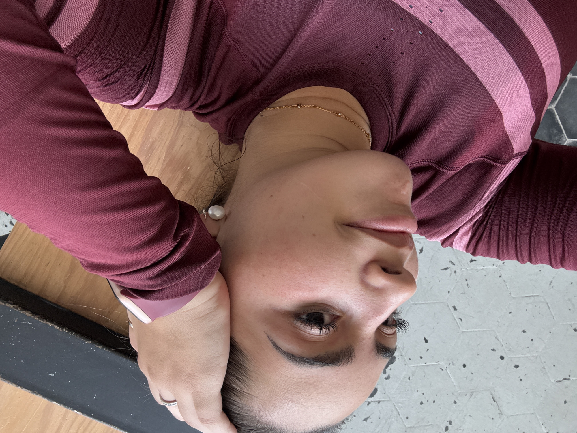 The image features a close-up, tilted, top-down view of a female subject, estimated to be in an indoor setting. The subject, with fair skin and dark hair pulled back, is wearing a long-sleeved, deep maroon top with lighter pink horizontal stripes across the chest and upper arm; the fabric displays subtle texture and small perforations near the shoulder. Her left hand, adorned with a silver-toned ring on the ring finger and a light-colored smartwatch band on the wrist, rests on top of her head. A prominent pearl earring is visible on her left ear, and a delicate gold chain necklace with small embellishments is around her neck. Her left eye is visible, featuring dark, elongated eyelashes and a well-defined eyebrow. Her gaze is directed slightly upwards and to the left within the frame. The background consists of two distinct areas: to the bottom and left, light grey hexagonal floor tiles with scattered dark speckles; to the top and right, a light brown wooden surface adjacent to a dark, rectangular object with a rough, lighter-colored edge. There is no visual information within the image to identify the specific city or location as Puebla, Mexico.