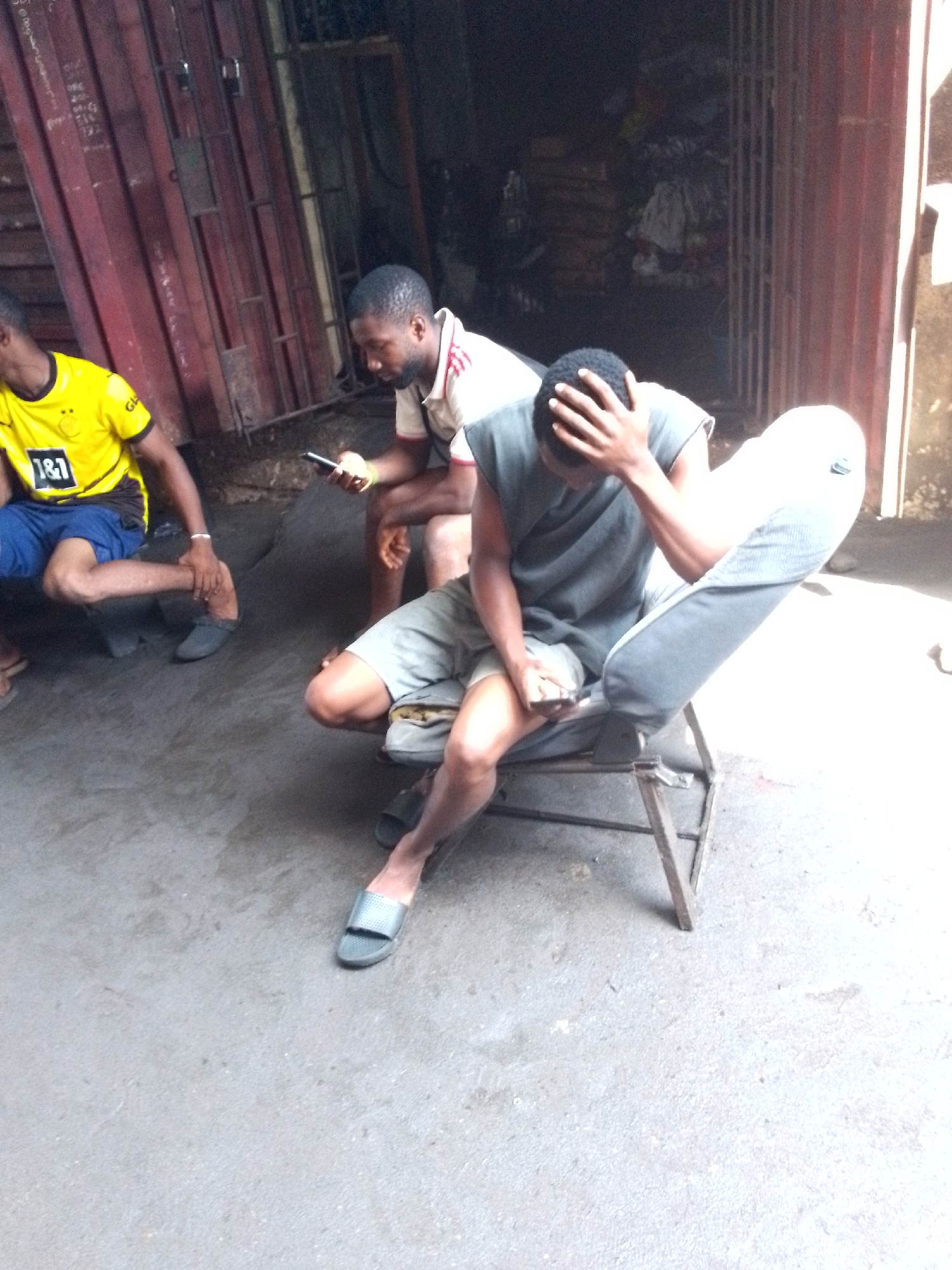 This daytime image features three men resting in what appears to be a semi-outdoor commercial or market setting. The ground is a light grey concrete or paved surface, with direct sunlight illuminating the right side of the frame and casting the left and background areas into shadow.
On the left, a man is partially visible, squatting or sitting on a low object. He is wearing a yellow and black football jersey, clearly displaying "1&1" and the logo of Borussia Dortmund (BVB), with "GLO" also partially visible. He is dressed in blue shorts and dark footwear.
In the center, a man with a short beard sits on a low, dark stool, looking down intently at a smartphone he holds in both hands. He is wearing a light-colored polo shirt with red stripes on the collar and placket, paired with dark shorts and dark slide-on slippers.
On the right, a third man is reclined on a simple, metal-framed chair with light grey fabric. He wears a dark grey sleeveless top and light grey shorts, with dark slide-on slippers. His left hand is raised, covering his face or head, while his right hand holds a small object, likely a phone, in his lap. His posture suggests weariness or deep thought.
The background includes a reddish-brown corrugated metal wall or gate on the left. In the center-right, a dark interior space, possibly a shop or storage area, is visible through an opening, containing indistinct stacked items. The scene captures a moment of respite, with at least two individuals engaged with their phones. The strong contrast in lighting suggests it is mid-day in an open or partially covered area.