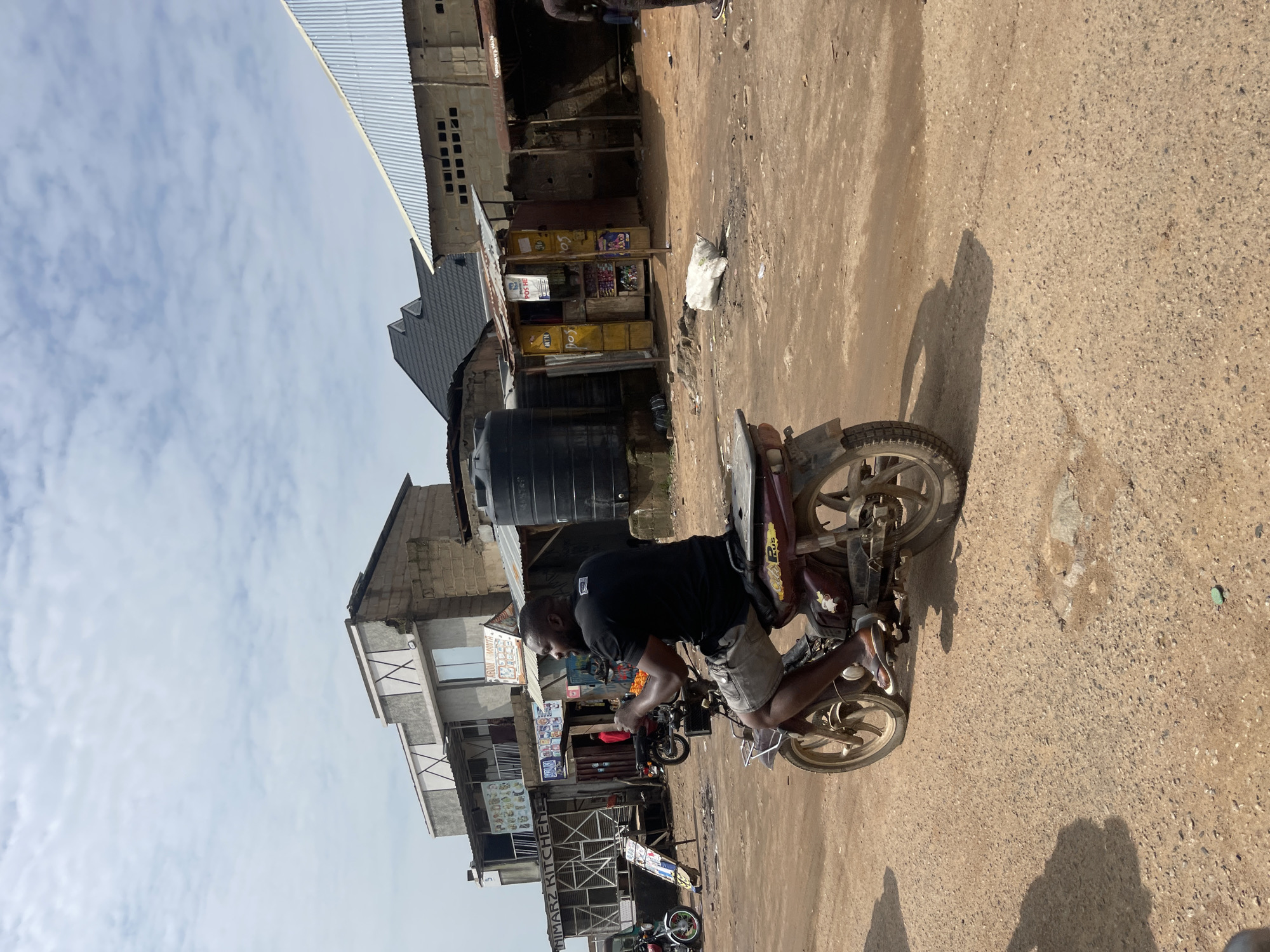 In Gwong, Nigeria, a man rides a motorcycle on a dirt road. The man is wearing a black t-shirt and grey shorts. The motorcycle is red and appears to be older. The road is unpaved and dusty, with several buildings and shops visible in the background. The buildings are made of concrete and corrugated metal, and some appear to be in disrepair. The sky is cloudy. There are other motorcycles and a large black water tank visible in the scene.