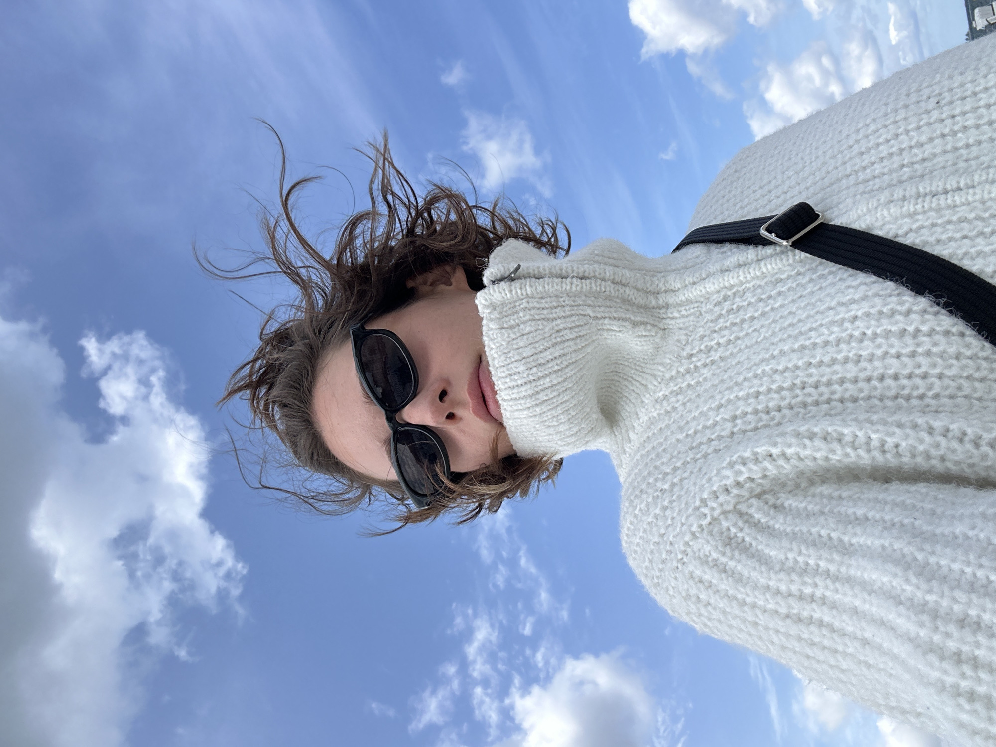 A low-angle, medium shot captures a person from the chest up, looking directly at the camera. The person is wearing a chunky, white, turtleneck sweater and black sunglasses. Their short, brown hair is windswept, suggesting an outdoor setting. The background is a bright blue sky with scattered white clouds. A black strap from a bag is visible across the person's shoulder. There are no discernible buildings or specific landmarks of Laboe, Germany visible in this image.