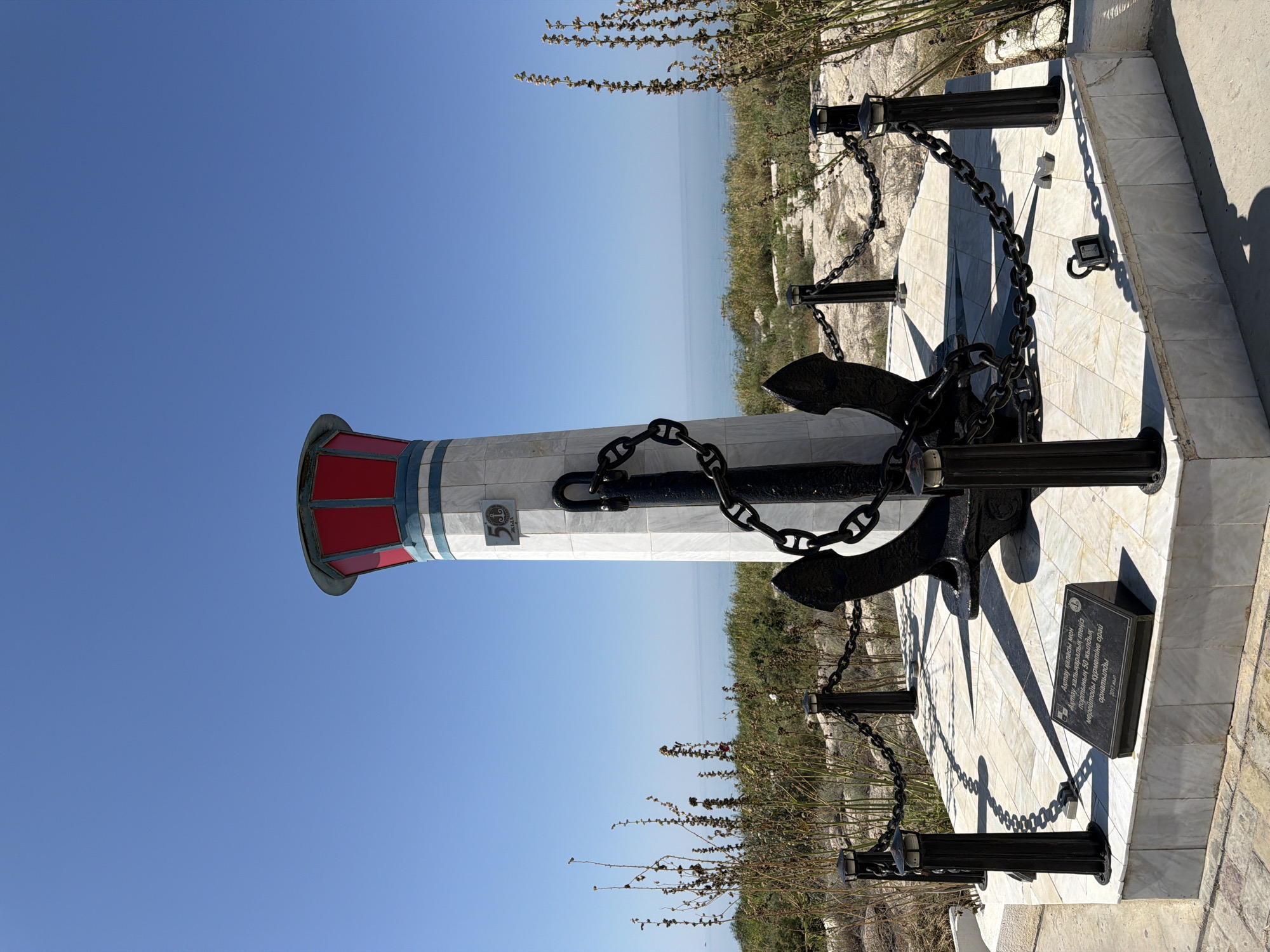 The image depicts a monument featuring a lighthouse-like structure with red accents at the top, and a large black anchor wrapped in chains at its base. The monument is situated on a circular, tiled platform, which is surrounded by a decorative chain fence supported by black posts with small lights. A plaque with text is visible at the front of the platform. The monument appears to be located outdoors, with dry vegetation and bushes in the foreground and background. In the distance, a calm body of water, likely the Caspian Sea, is visible under a clear blue sky. The monument commemorates the 50th anniversary of the port of Aktau, Kazakhstan.
