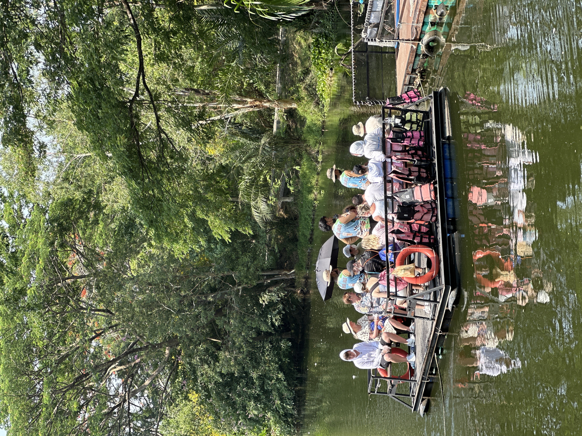 A daytime shot in Bang Phra Subdistrict, Thailand, displays a long, rectangular boat populated with approximately 15 individuals. The boat is positioned on a murky, green body of water, which reflects elements of the boat and its occupants. Passengers are seated, most appearing to be wearing light-colored attire and some are wearing hats and life jackets. An open black umbrella is visible among the passengers. The boat has a black metal frame and some orange life rings. Along the perimeter of the waterway, trees with dense green foliage are observed. A wooden structure or dock with attached tires is at the end of the boat.