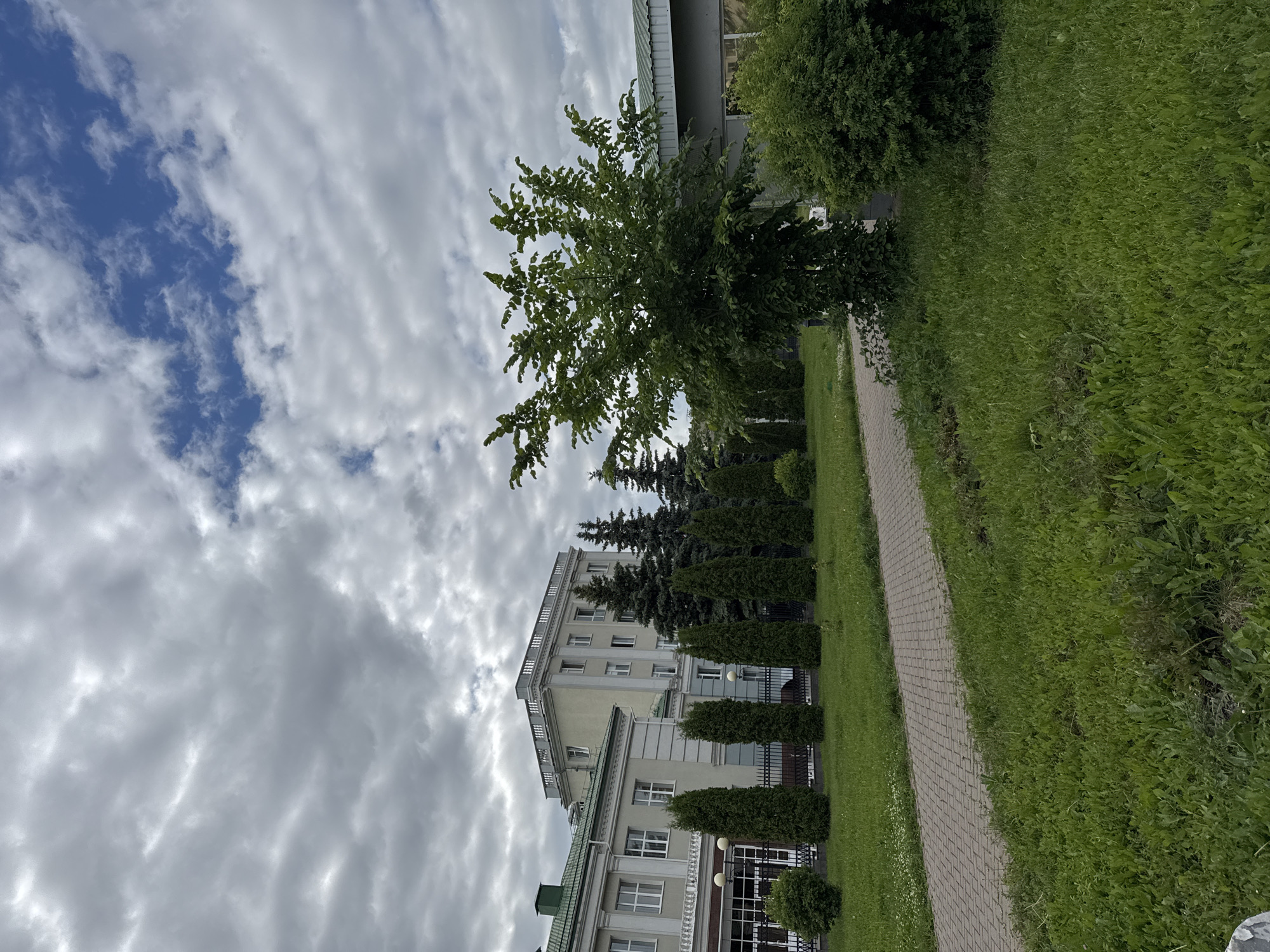The image depicts an outdoor scene with a building and landscaping. The building appears to be a multi-story structure with a light-colored facade and a green roof. In front of the building, there is a paved walkway and a lawn with several evergreen trees and bushes. The sky is partly cloudy. The location is the Izhevsk State Medical Academy, located at Lenina Street, 1, Izhevsk, Udmurt Republic, Russia.