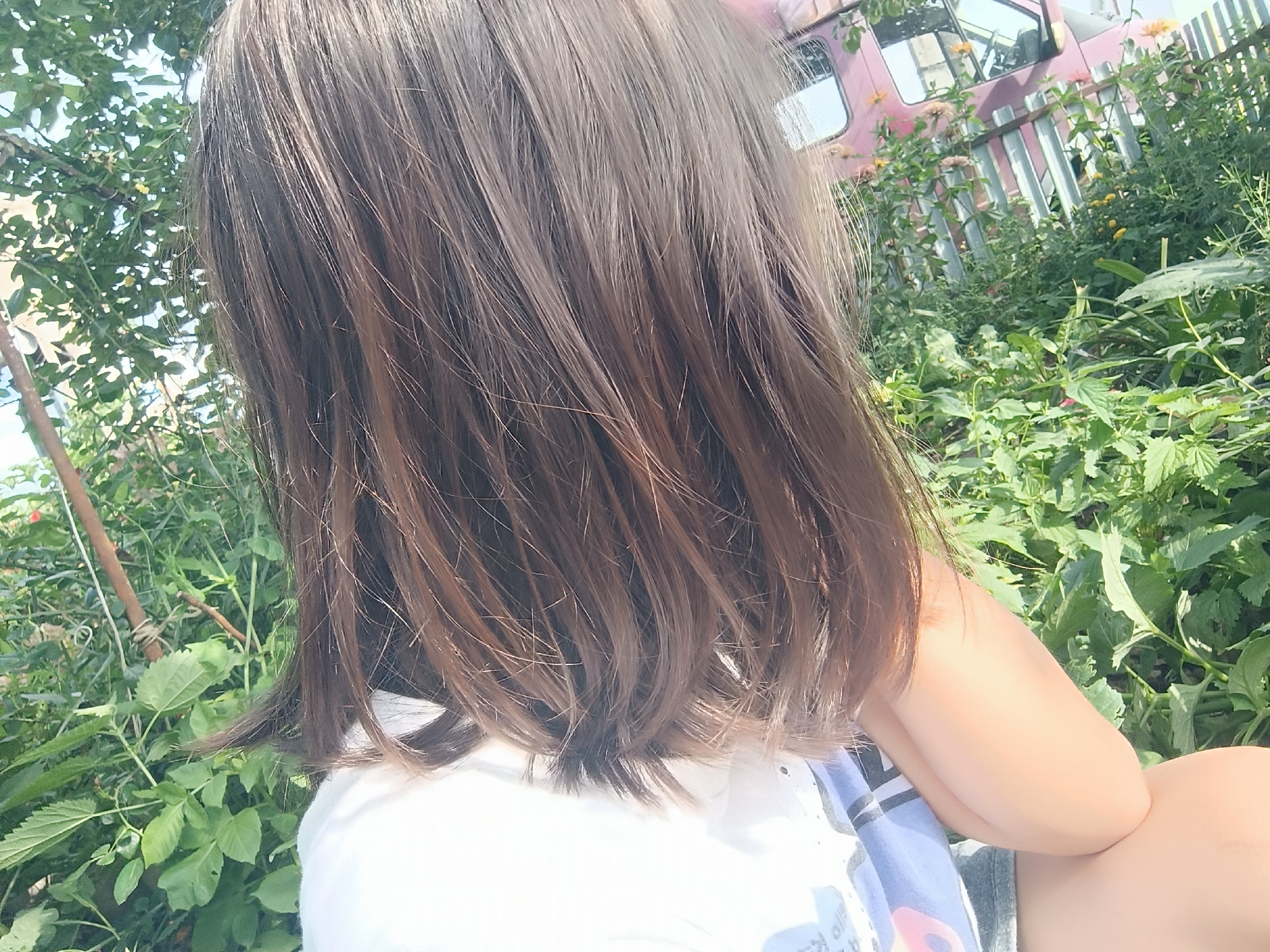 The image shows a person from behind, with their head turned to the right. They have medium-length brown hair. They are wearing a white t-shirt with a design on it. The person is outdoors, with lush green foliage and plants surrounding them. In the background, there is a light pink structure that appears to be a vehicle or part of a building, with a wooden fence in front of it. There are also some small yellow flowers visible. The lighting suggests it is daytime, possibly late afternoon due to the warm tones. The scene is set in an outdoor environment, likely a garden or a yard. There is no information about the specific location within the city of Karsha, Russia, in the image itself.