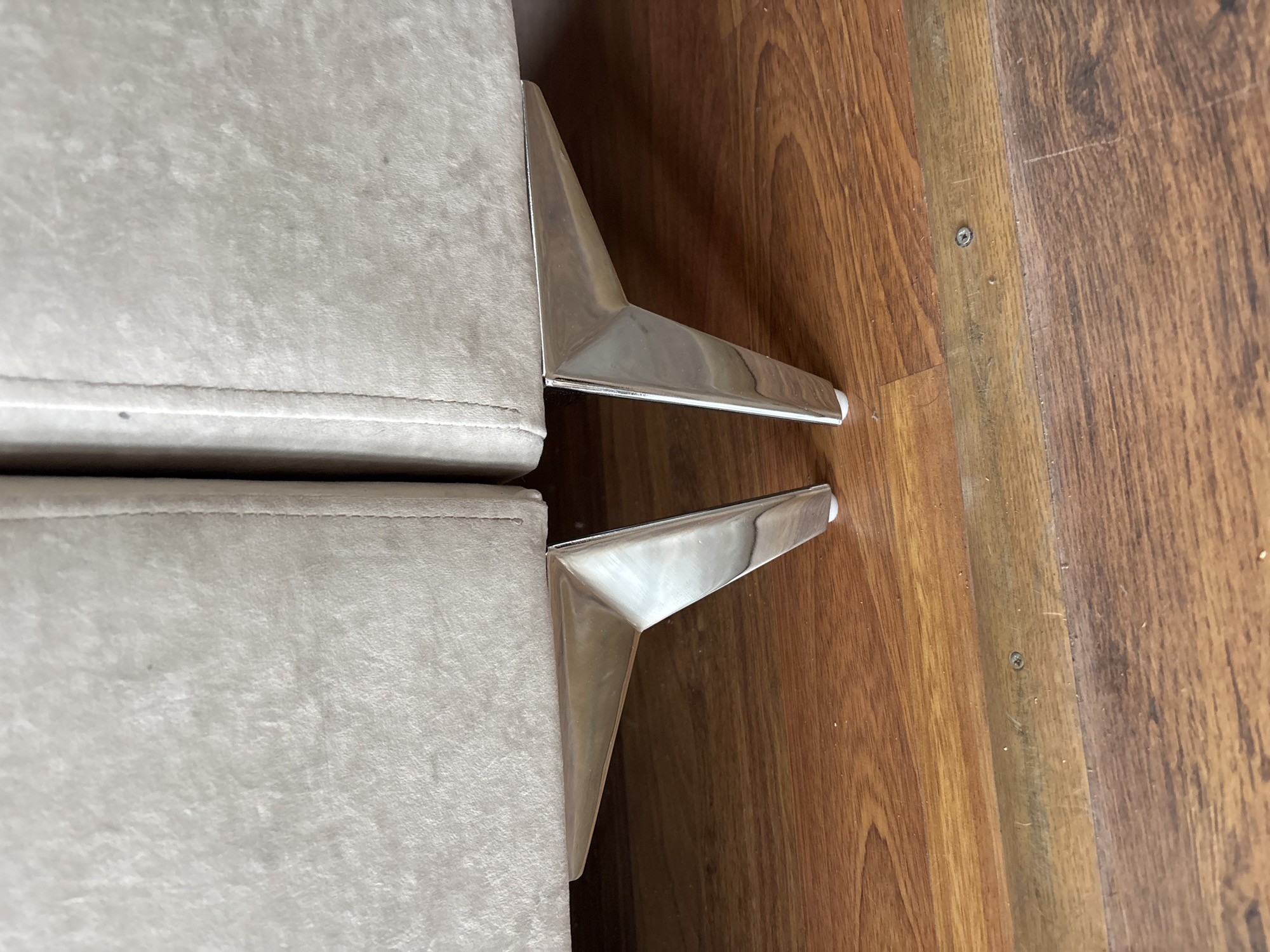 This image shows the bottom portion of a light-colored sofa with two metallic, triangular legs resting on a wooden floor. The floor appears to be made of planks of varying shades of brown, with a darker strip of wood running horizontally across the lower part of the frame, featuring a visible screw. The sofa fabric is a plush, light beige color. The legs are angled and appear to be made of a polished metal, reflecting the surrounding light. The scene is indoors, and there are no people visible. The location is not specified.