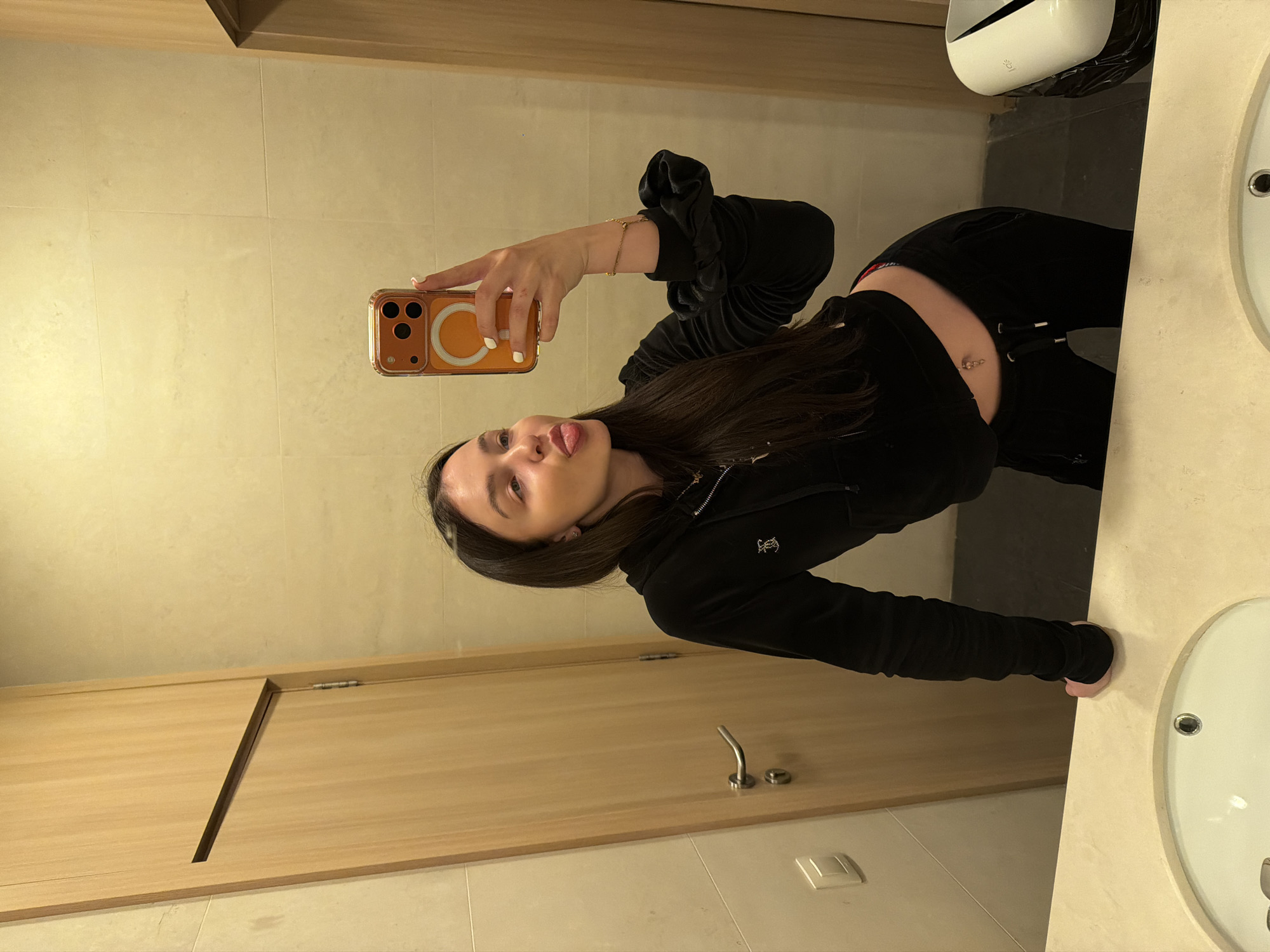 A young woman is captured in a mirror selfie taken in what appears to be a modern restroom, likely in Sofia, Bulgaria. She has long dark hair and is wearing a dark, possibly velvet, zip-up top with a "Juicy Couture" logo (a crown above "JC") on the left chest, paired with dark trousers. Her midriff is exposed, revealing a belly button piercing.
She holds an orange-cased smartphone, possibly an iPhone with a visible MagSafe ring, in her right hand, positioned to capture her reflection. Her left arm is visible, adorned with a delicate gold-colored bracelet, and her fingernails are painted white. The woman's head is slightly tilted, and she is playfully sticking her tongue out, conveying a lighthearted expression.
The background shows light-colored tiled walls and wooden paneling, including a door with a distinctive diagonal cutout. A chrome handle and a white light switch are visible on the wooden surface. To the right, a white sink with a chrome drain is prominent, and above it, a white hand dryer or dispenser is mounted, partially obscuring a black trash bag. The lighting is bright and even, consistent with an indoor, artificially lit environment. The overall impression is a casual, playful moment captured in a functional, clean setting.