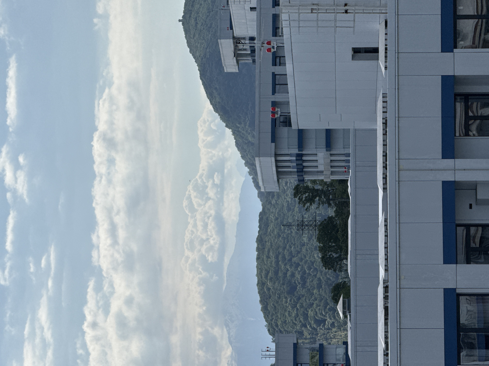 Here is a description of the image:
The image displays a view combining natural and urban elements, possibly in the city of Sochi, Russia. The sky is visible on the left, filled with a pattern of scattered clouds. Adjacent to the sky are mountains covered with dense green forest vegetation.
A modern building with multiple floors and windows is visible on the right side of the image. The building appears to have a neutral color scheme, with blue accents on the facade.
The overall atmosphere suggests a location where modern architecture coexists with the natural landscape, possibly in an urban setting with access to mountainous terrain.