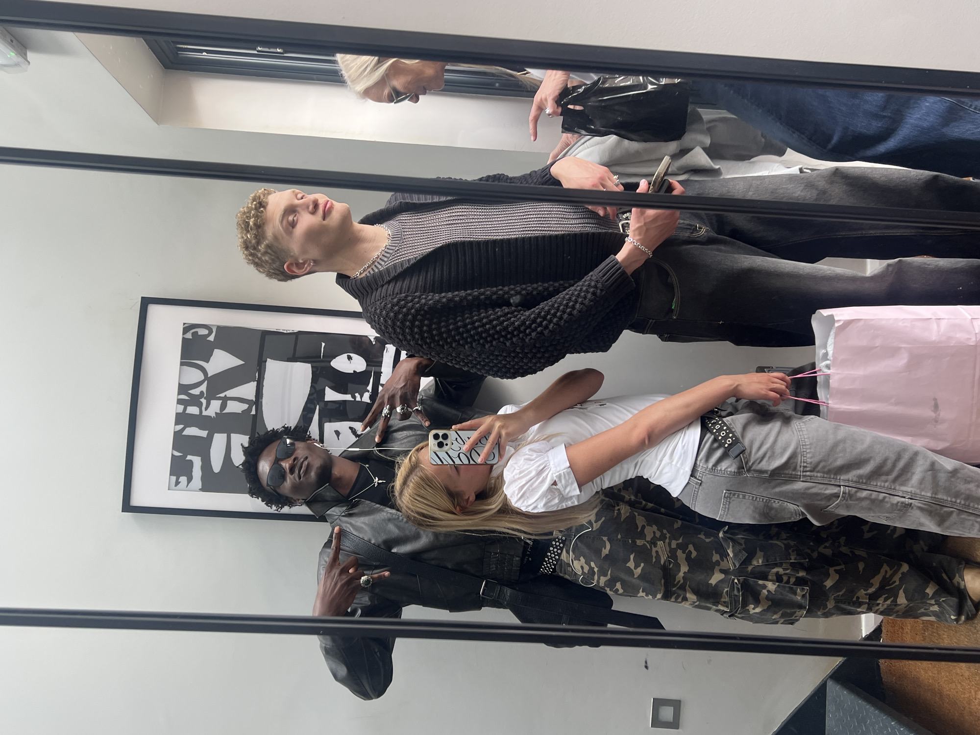 A mirror selfie of four people in what appears to be a room or retail space. The location is possibly in London, United Kingdom. In the reflection, a light-skinned male with curly blond hair looks slightly up, wearing a black knit cardigan over a gray and white striped shirt, and black jeans. Above him, a woman with blond hair and sunglasses is partially visible.
Below the man, a dark-skinned male with short curly black hair wears sunglasses, a black t-shirt, and a black leather jacket, making a peace sign with his left hand.
A light-skinned woman with long blond hair is holding a phone, presumably taking the picture. She wears a white shirt and gray jeans with a studded belt. There is a pink shopping bag beside her.
Behind the figures in the reflection, there is a black and white framed poster on the wall. The room appears to have a neutral color scheme, with a white wall. The floor is partially visible at the bottom of the mirror, appearing to be carpeted. The mirror is surrounded by a black frame.