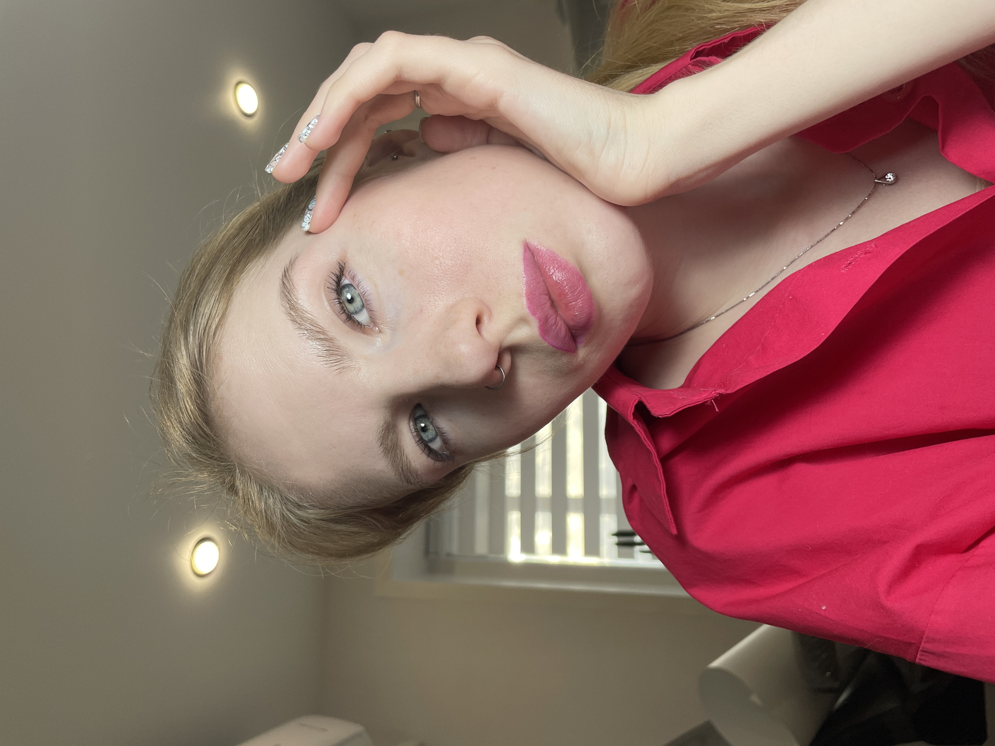 A close-up portrait, oriented vertically, captures a young person with light blonde hair and striking blue-green eyes, looking directly at the camera. Their head is tilted, and they wear a bright red collared shirt. Their lips are painted with a soft pink lipstick, and a silver hoop nose piercing adorns their left nostril. The person's left hand is raised, with fingers gently resting on their forehead and temple. Their long fingernails feature a light, possibly glittery or patterned, manicure. A delicate silver chain necklace with a small, spherical pendant is visible around their neck.
The setting appears to be an indoor room with light-colored walls. Two circular, warm-toned light fixtures are visible on the wall in the upper left background. To the right, a window with horizontal blinds can be seen, allowing light to filter through and create vertical patterns. The overall lighting seems to be a combination of artificial and natural light. The person's expression is neutral and direct. No visible text is present in the image. The image is set in Izhevsk, Russia.