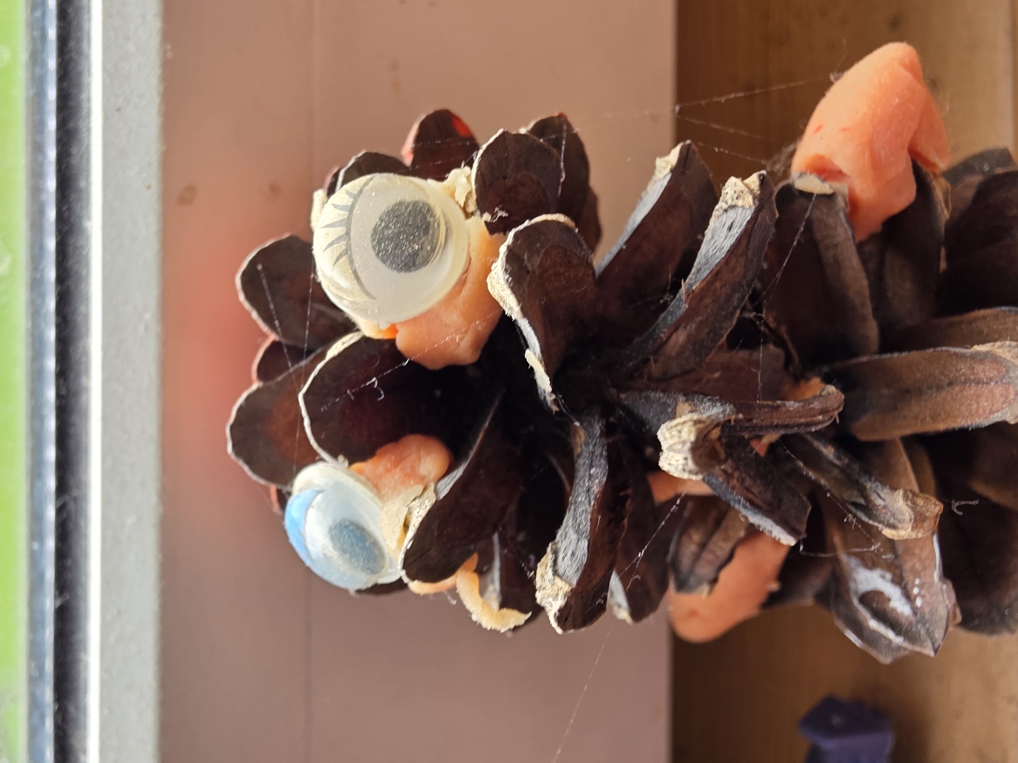 An upward-oriented, close-up shot features a decorated pinecone, likely a craft item or decoration. The pinecone itself is a rich dark brown with visible, textured scales. It is adorned with two googly eyes: one on the upper side is white with a grey pupil and delicate drawn-on eyelashes, while the other, positioned slightly lower, is blue with a darker pupil. Interspersed between the pinecone's scales are several pieces of soft, orange-colored craft material, possibly foam or clay. Fine, delicate cobwebs are visible across the surface of the pinecone and extending faintly into the background, suggesting it has been stationary for a period.
The background on the left side of the image consists of blurry vertical elements in shades of green and grey, which could be part of a window frame or a nearby structural element. The primary background on the right is a plain, muted light orange or peach-colored wall or surface, exhibiting a few small, faint brown spots. The overall lighting is bright and even, indicating either ample ambient light or direct illumination. There is no specific activity or event depicted; the image is a still-life photograph of the crafted object. The googly eyes imbue the pinecone with a whimsical, personified character. No text is visible anywhere in the image. The location context provided is Vidnoye, Russia.