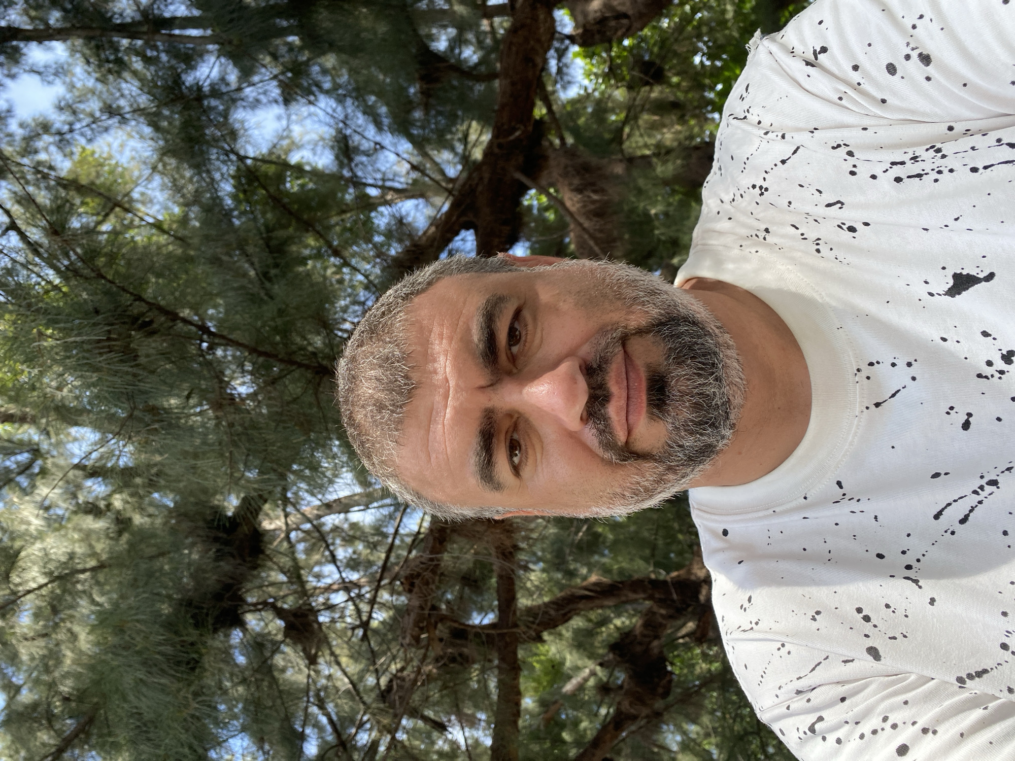 A middle-aged man with graying hair and a beard is photographed from the chest up, outdoors, with trees and a blue sky in the background. He is wearing a white t-shirt with black paint splatters. The man is looking directly at the camera with a neutral expression. The image appears to be a selfie taken in a park or forested area. The location is not identifiable as Nha Trang, Vietnam.