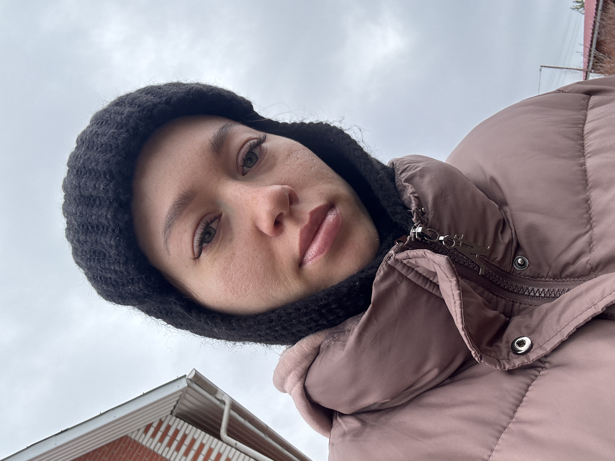 A low-angle selfie depicts a young woman wearing a black knit hood and a mauve-colored puffy jacket. She is facing the camera with a neutral expression, her face slightly angled. Above her, a gray, overcast sky is visible. In the lower left corner, the edge of a brick building with a white roofline is present. A faint powerline crosses the upper right area of the image. The photo was likely taken outdoors in a residential area in Ферма, Russia.