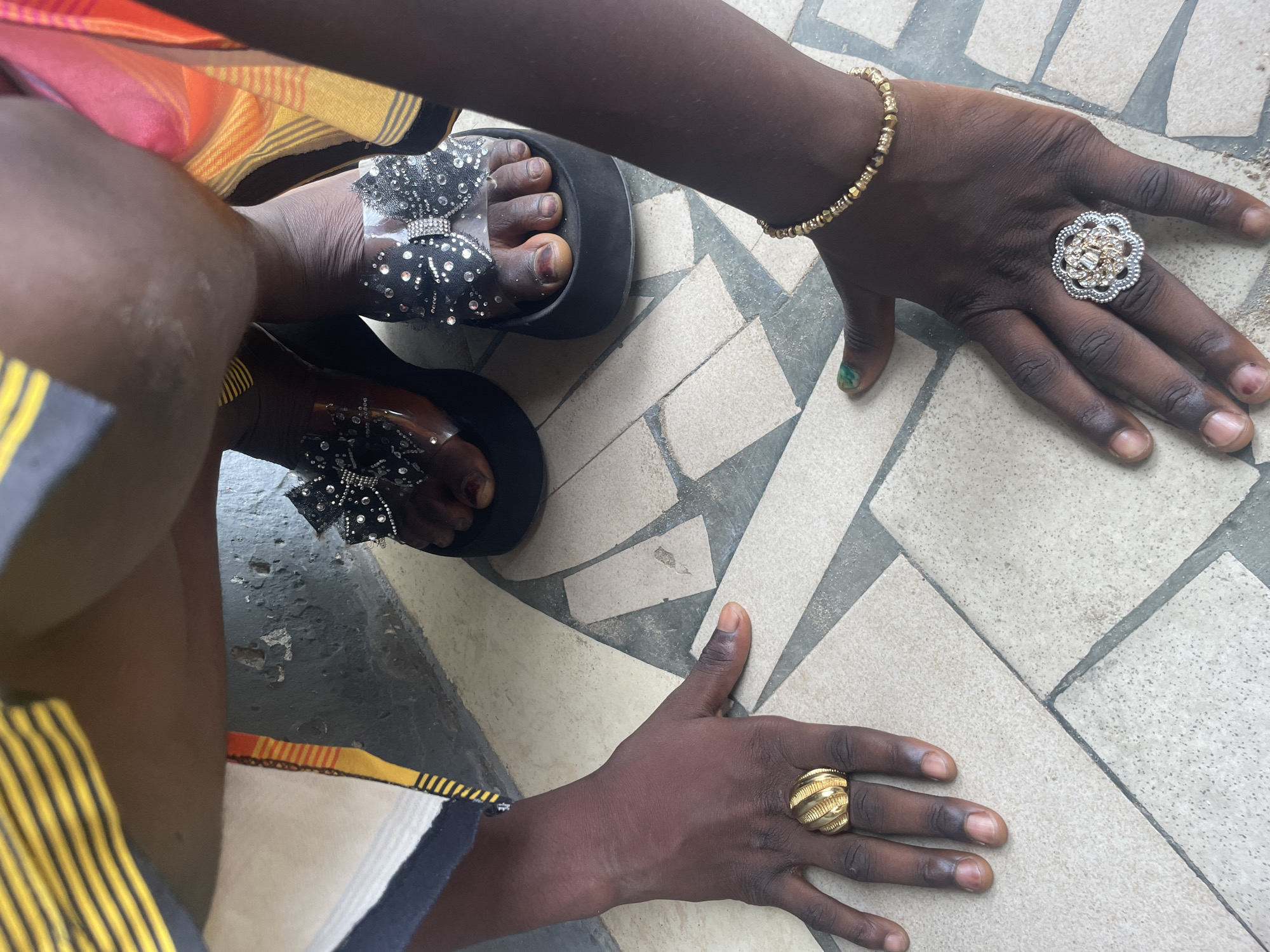 The image provides an overhead view of a person's hands and feet, showcasing various accessories against a tiled floor, likely in Badagry, Nigeria.
The subject has dark skin. The right hand displays a gold-colored beaded bracelet on the wrist and a large, ornate silver-toned ring with a floral, sparkling design on the middle finger. A faint greenish hue is visible on the pinky fingernail. The left hand wears a prominent, spherical gold-toned ring with a ribbed texture on the ring finger. The fingernails on both hands appear natural or unpolished.
The feet are wearing fashionable black platform sandals. These sandals feature clear, transparent straps across the toes and instep, adorned with elaborate black bow embellishments covered in rhinestones. The toenails are natural and unpolished.
Portions of clothing are visible: a vibrant fabric with stripes in orange, yellow, and black is seen on the upper left, while a striped fabric in yellow, black, and white is on the lower left.
The setting is a tiled floor composed of light beige or off-white rectangular and triangular tiles separated by dark grey grout lines, arranged in a geometric pattern. The lighting suggests a daytime setting, possibly indoors or in a shaded area. There is no explicit activity taking place, but the arrangement implies a deliberate pose to highlight the jewelry and footwear. No text is visible in the image.