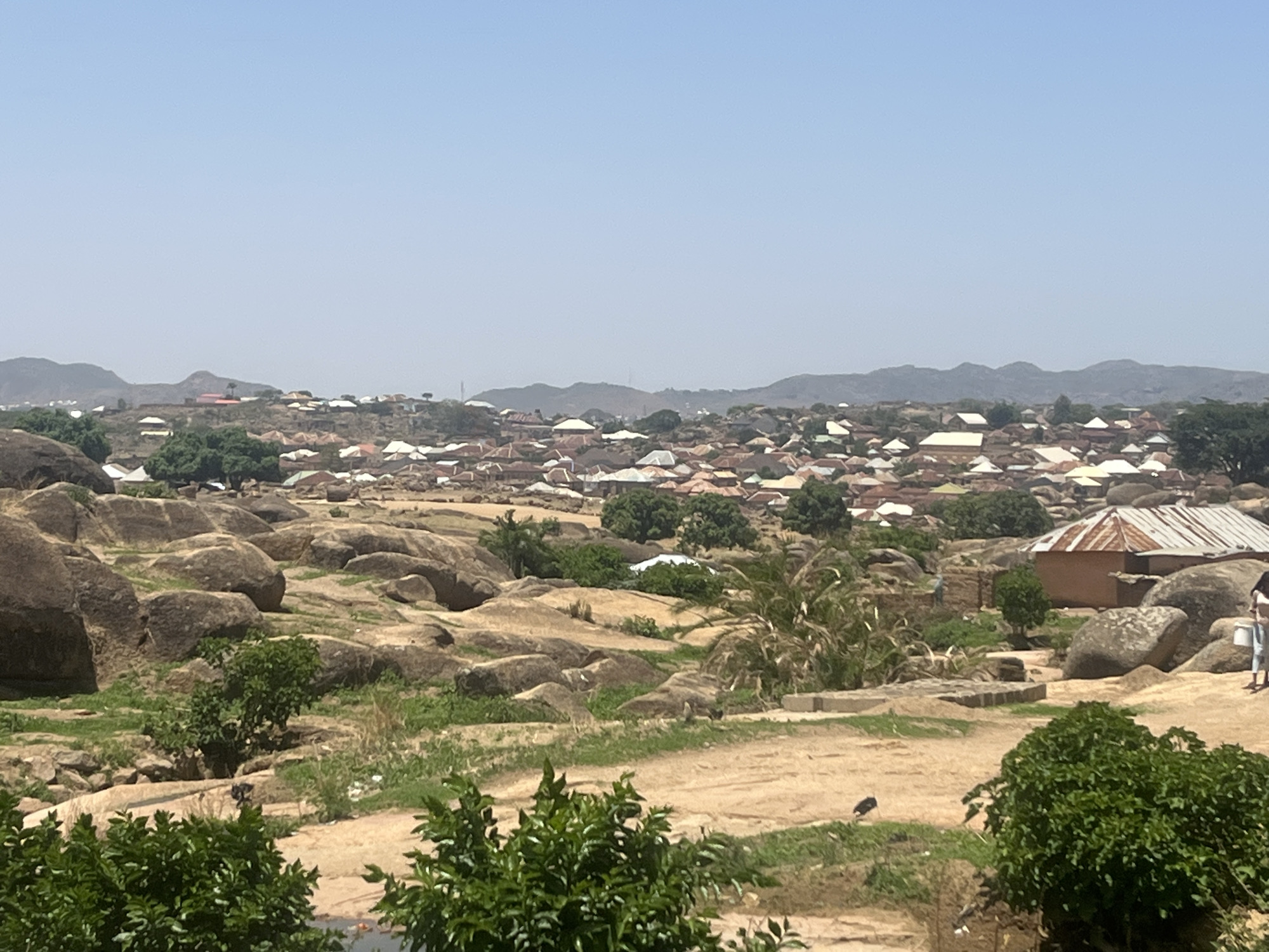 The image depicts a landscape in Jos, Nigeria, characterized by large, rounded rocks scattered across the terrain. Vegetation includes green trees and scrub bushes. A cluster of buildings with light-colored roofs occupies the middle ground, merging with the landscape. In the distance, mountain ranges are visible under a clear, blue sky. A building with a metallic roof stands out amid the rocks. A person is present towards the right with a white container or pail. The foreground consists of dirt and low-lying greenery.