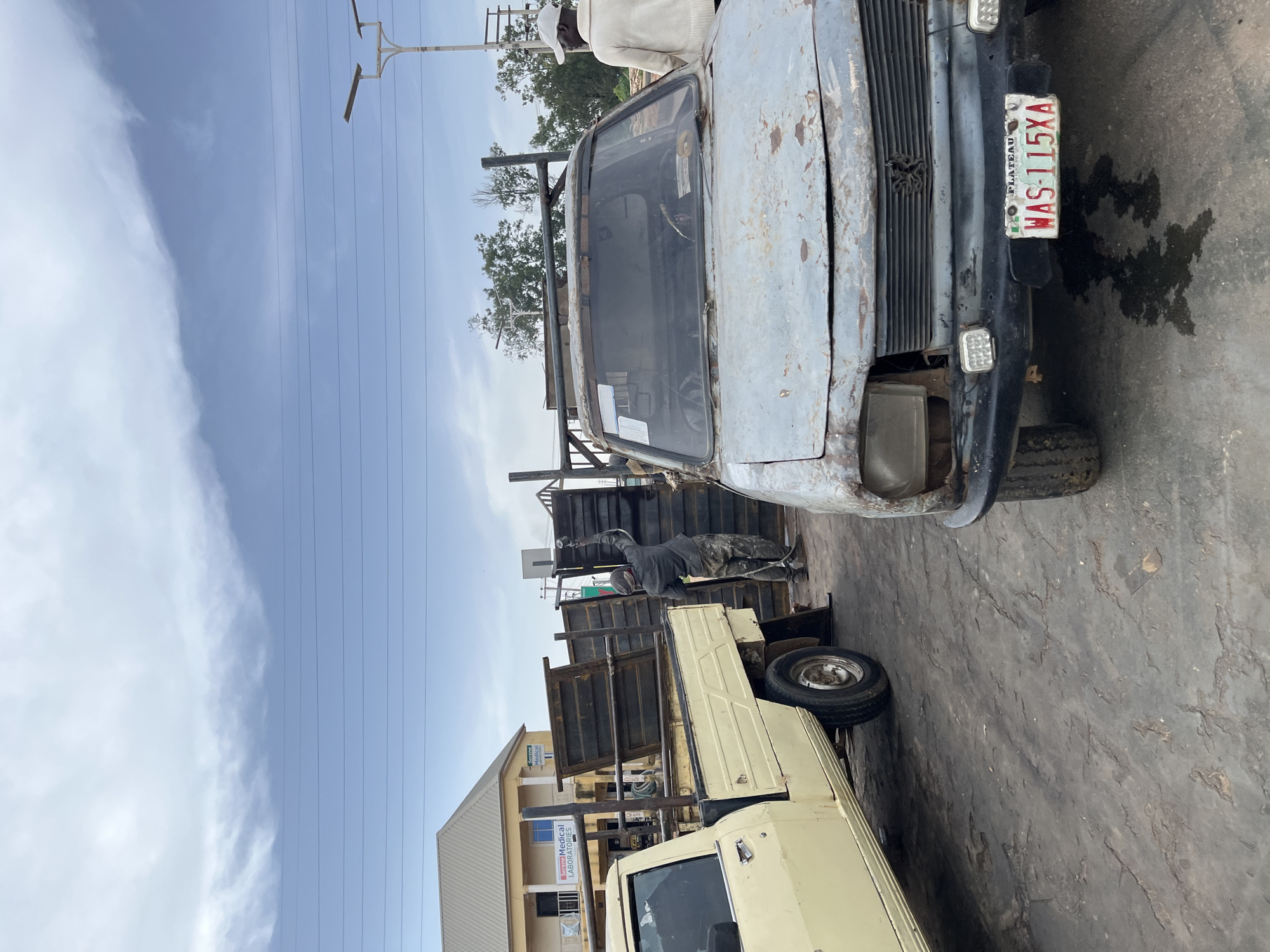 The image depicts an outdoor scene in Gura Top, Nigeria, featuring several vehicles and a building. The primary focus is on two pickup trucks, one old and rusty with a light blue exterior, and another newer, light yellow truck behind it. The rusty truck has a visible license plate that reads "WAS-115XA" and "PLATEAU". A person wearing a white cap and a cream-colored sweater is standing near the front of the rusty truck, looking towards it.
In the background, a building with a light beige facade and blue trim is visible. Signs on the building include "Supreme Medical Laboratories" and another that is partially obscured. Several power lines stretch across the sky, suggesting the presence of electrical infrastructure. The sky is cloudy, indicating a possibly overcast or partly cloudy day. The ground appears to be asphalt or concrete. The overall scene suggests a street or a yard where vehicles are parked, possibly a marketplace or a mechanic's workshop. The lighting suggests it is daytime, likely late morning or early afternoon. The condition of the older truck suggests it might be disused or awaiting repair.