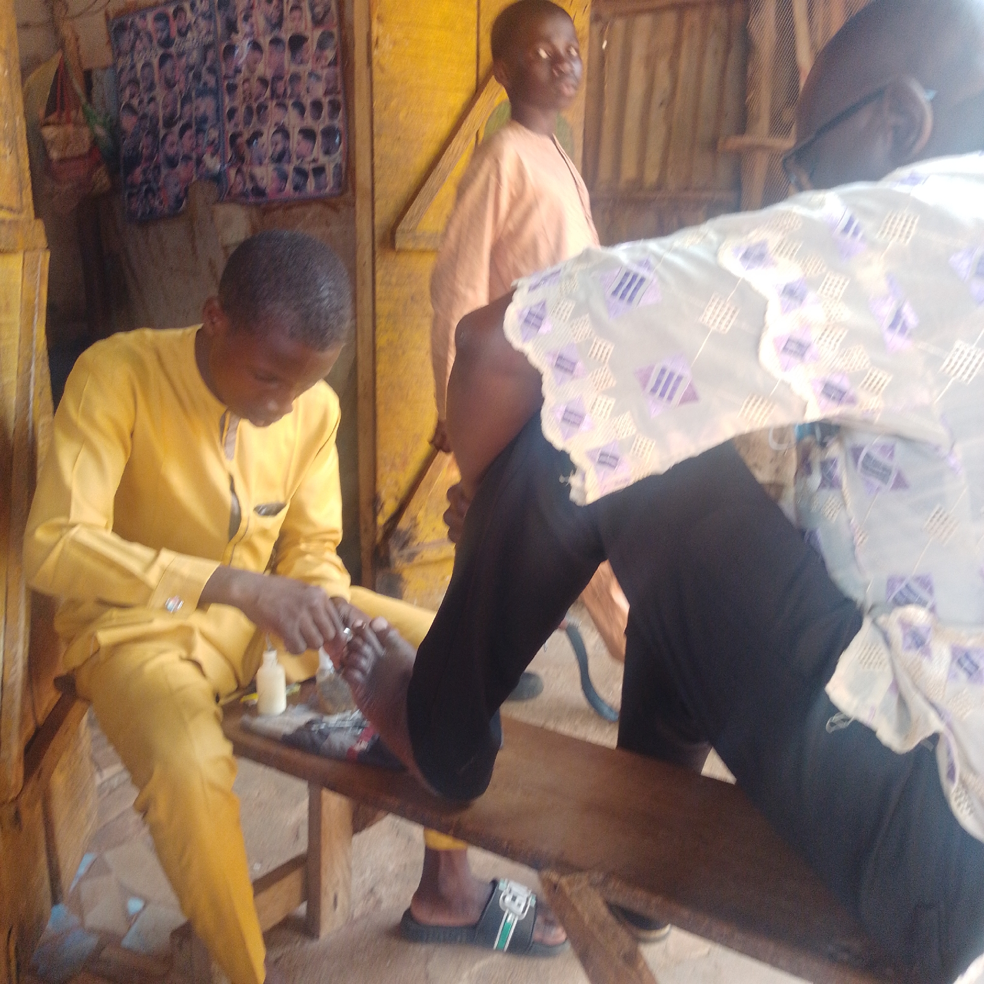 In Gombe, Nigeria, a young individual in a yellow outfit is seated on a wooden bench, meticulously performing a pedicure on another person's foot. The recipient of the pedicure is wearing black trousers and a patterned shirt, with their foot resting on the bench. A third person, also in a pale orange garment, stands behind, observing the procedure. The setting appears to be an open-air establishment, with wooden structures and possibly posters of hairstyles visible on the wall in the background. A small white bottle, likely containing nail polish or lotion, is positioned on the bench near the foot being worked on. The ground is dirt.