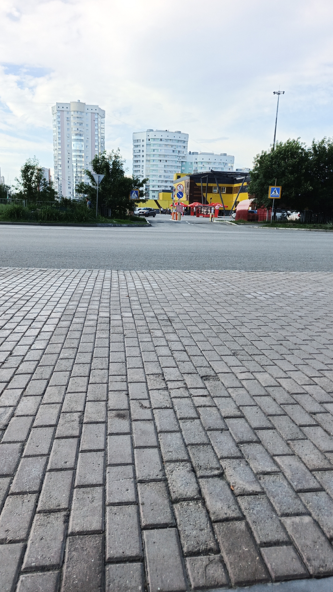 The image depicts a street scene in Yekaterinburg, Russia, with a foreground of grey interlocking pavers leading to a paved road. Beyond the road, there are several multi-story residential buildings with white and light-colored facades and numerous windows. In the mid-ground, a yellow building with a dark roof structure is visible, featuring red umbrellas and what appear to be food stalls or outdoor seating areas. Several individuals, dressed in bright yellow, are gathered near these stalls. Traffic signs, including a pedestrian crossing symbol, are also present. Trees with green foliage line the sides of the road and separate the buildings from the street. The sky is overcast with scattered clouds. The perspective suggests the photograph was taken from a low angle, looking across the street towards the buildings and commercial area.